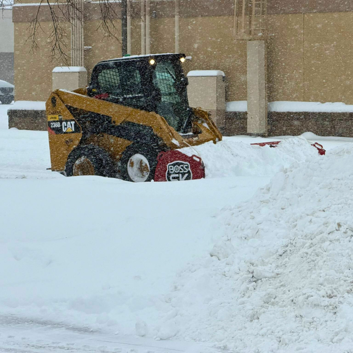 A yellow CAT skid-steer loader equipped with a snow plow attachment clearing snow in an urban area during snowfall.
