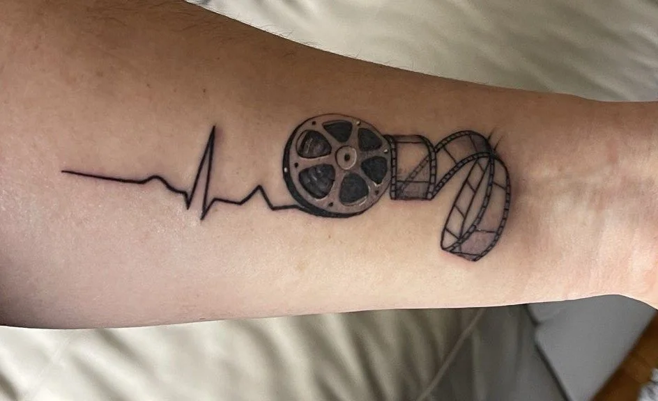 Close-up of an inner forearm with a black-and-grey tattoo showing a heartbeat line flowing into a vintage film reel, with a strip of film curling outward.