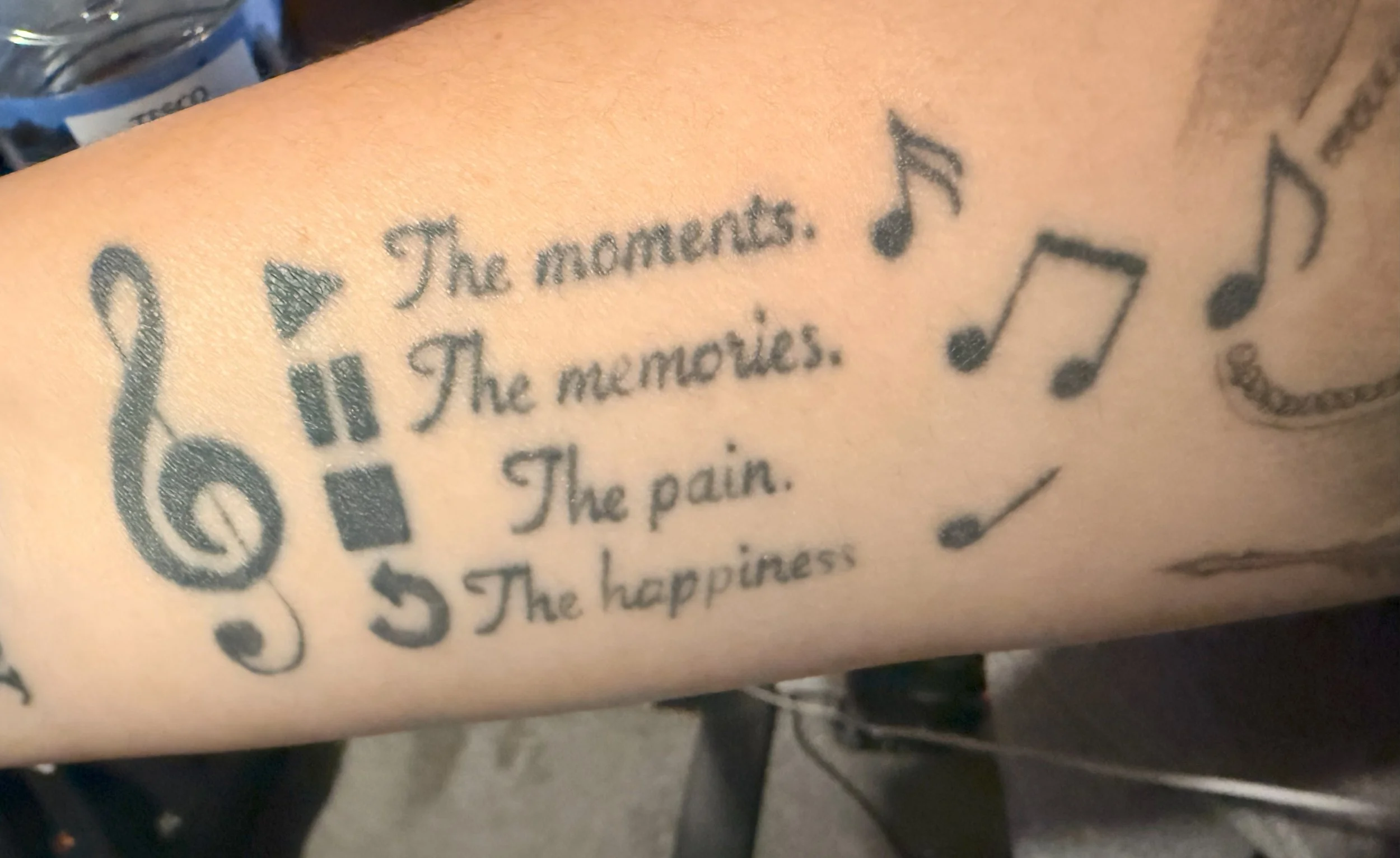 Close-up of a forearm tattoo combining musical symbols with media controls. A treble clef sits beside play, pause, stop and replay icons, alongside the words “The moments. The memories. The pain. The happiness.” Musical notes surround the design.