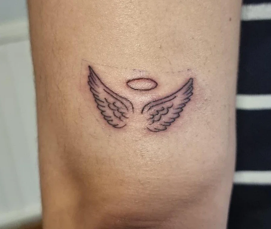 Close-up of an arm with a small fine-line tattoo of two angel wings beneath a simple halo, positioned just above the elbow.