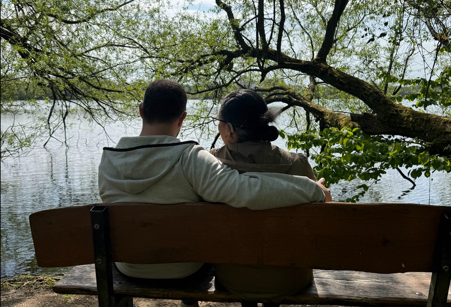 Photograph of two people sitting side by side on a wooden bench, facing a calm lake. One has their arm around the other as they look out across the water.