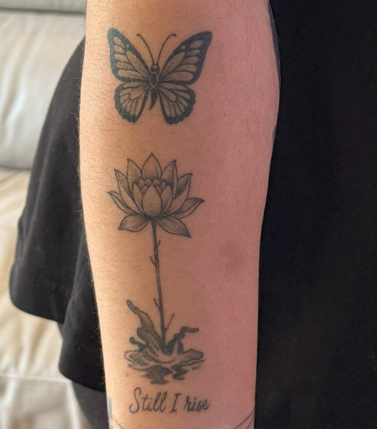 Photograph of a forearm showing a tattoo of a butterfly above a lotus flower growing from water, with the words “Still I rise” written below. The image is natural and closely framed, showing the detail of the design on the skin.