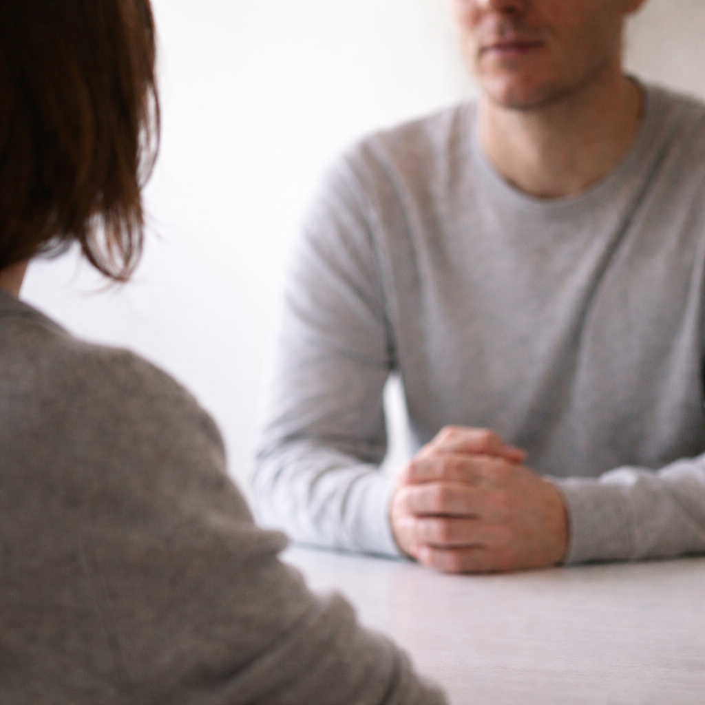 Soft, muted photograph of two people sitting across from each other at a table in a blurred indoor setting.
