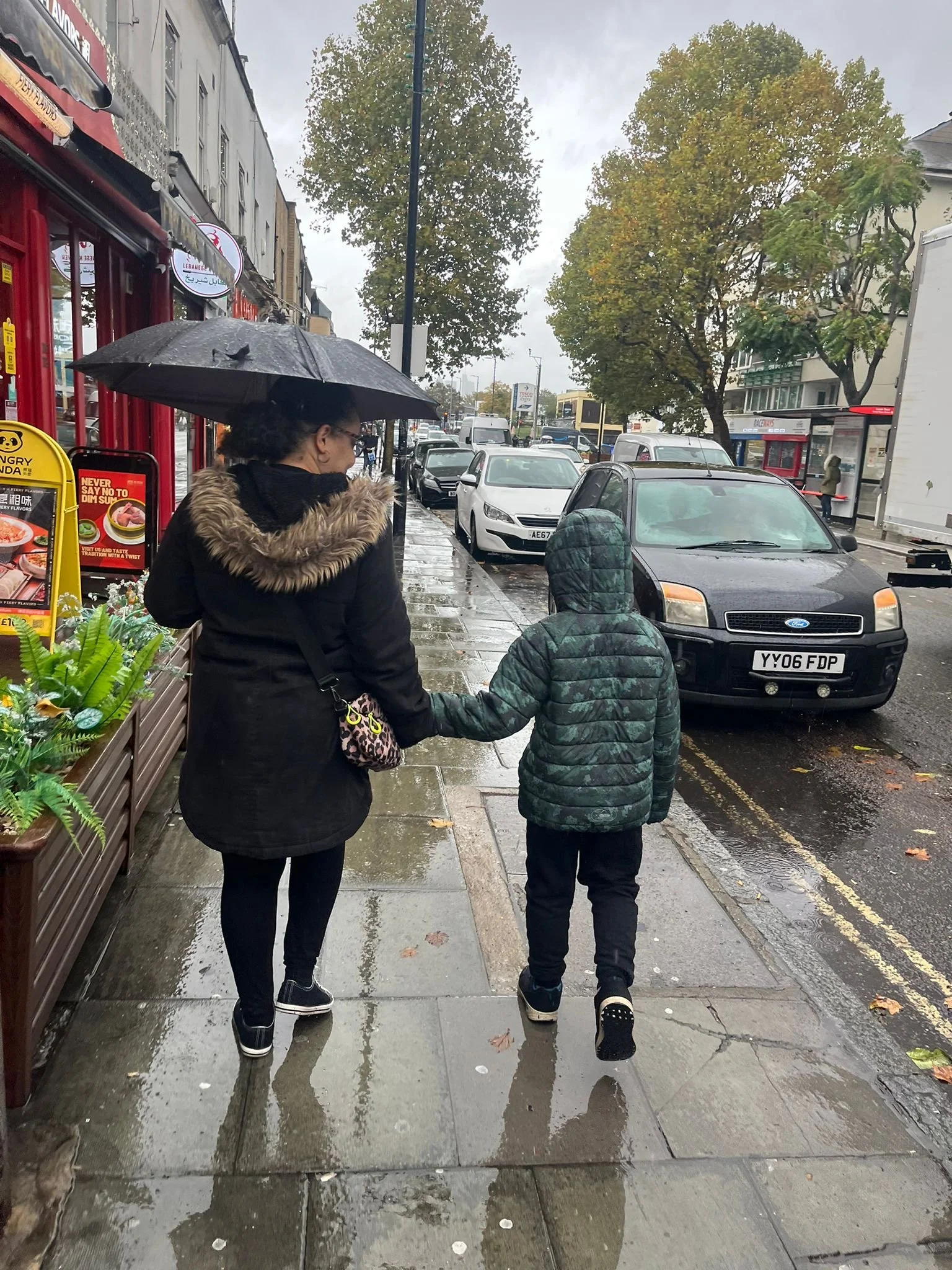 Photograph of an adult and a child walking hand in hand along a wet pavement on a rainy street, viewed from behind. The adult holds an umbrella while the child walks beside them in a hooded jacket.