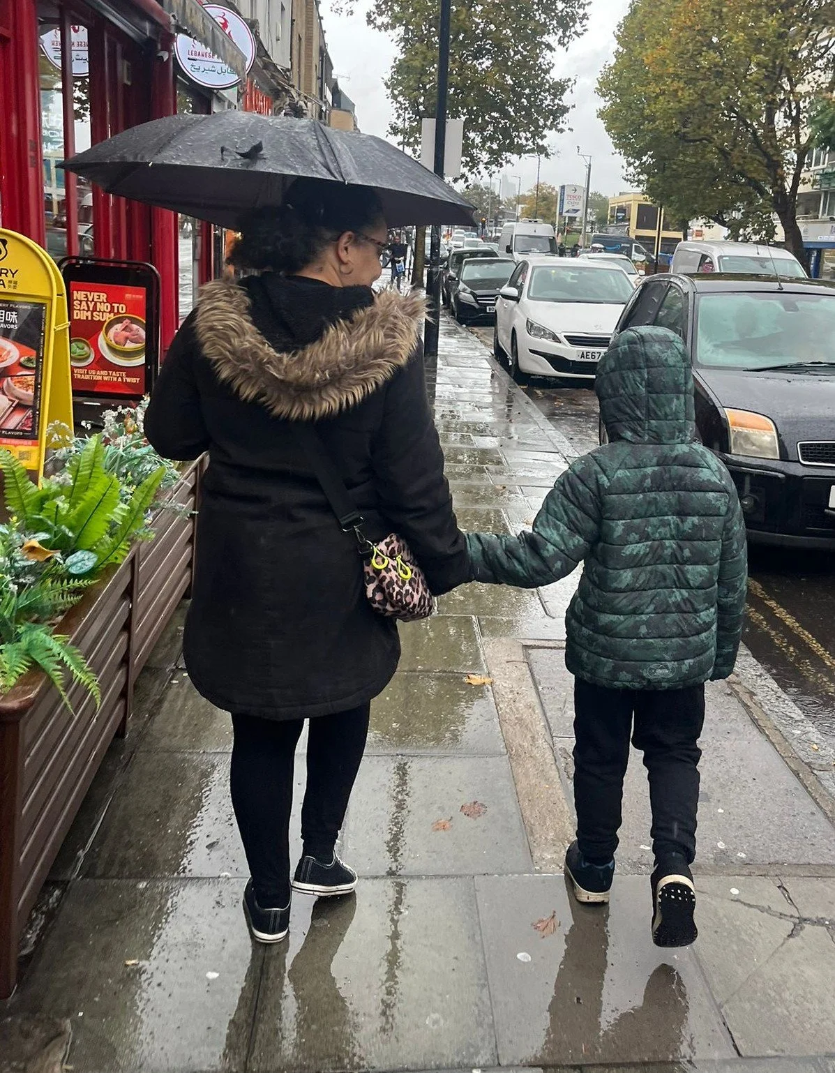 Photograph of an adult and a child walking hand in hand along a wet pavement on a rainy street, viewed from behind. The adult holds an umbrella while the child walks beside them in a hooded jacket.