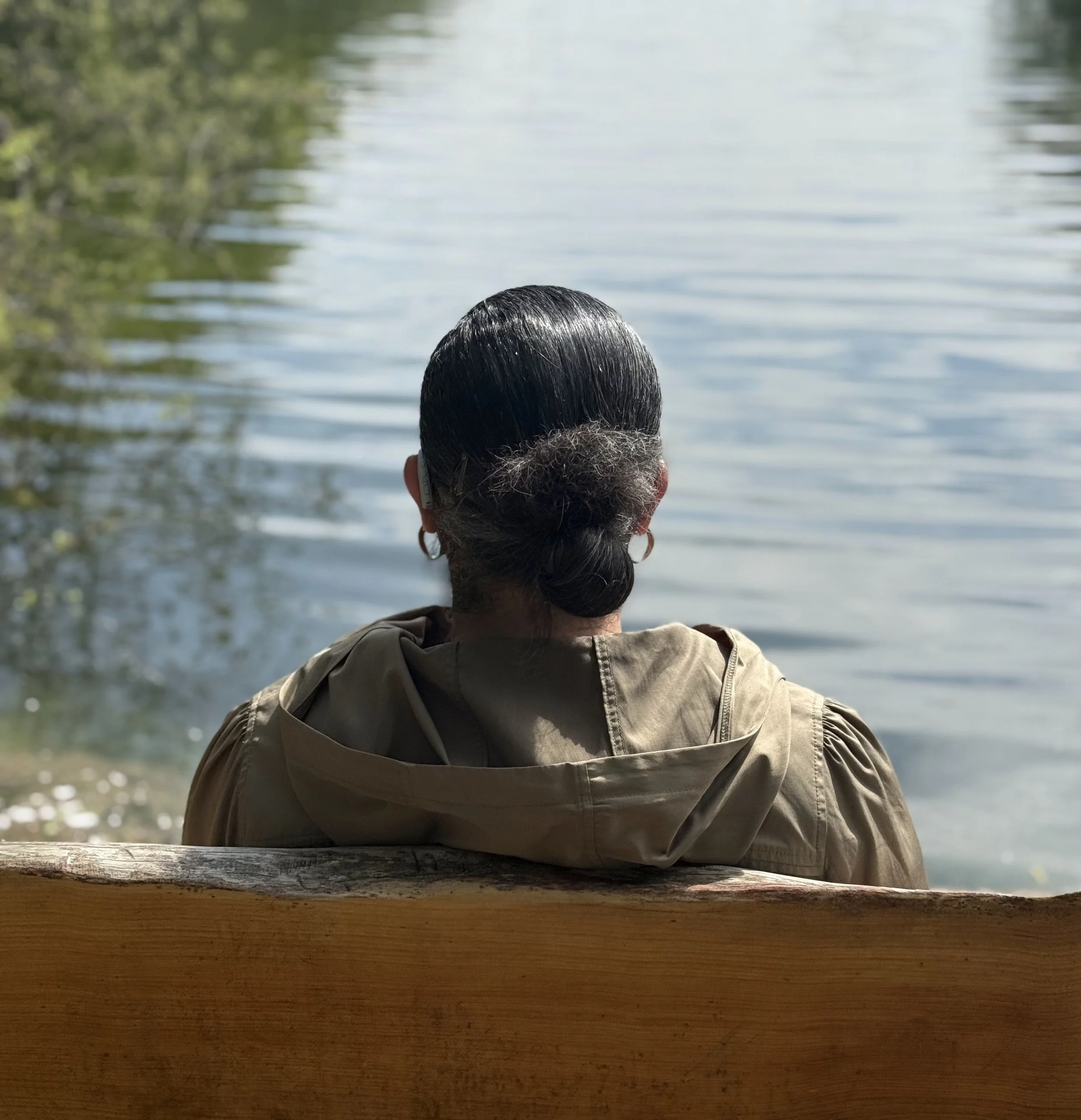 Photograph of a person sitting on a wooden bench facing a calm lake, viewed from behind. Trees frame the water on both sides, and the surface of the lake is still with soft ripples.