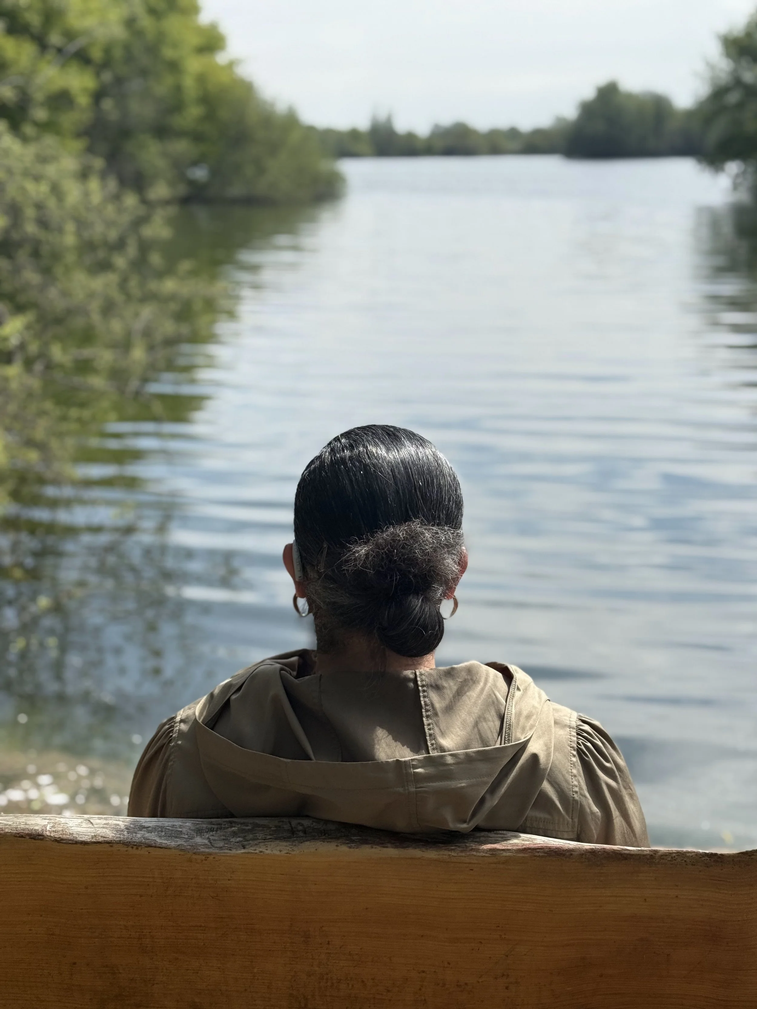 Photograph of a person sitting on a wooden bench facing a calm lake, viewed from behind. Trees frame the water on both sides, and the surface of the lake is still with soft ripples.