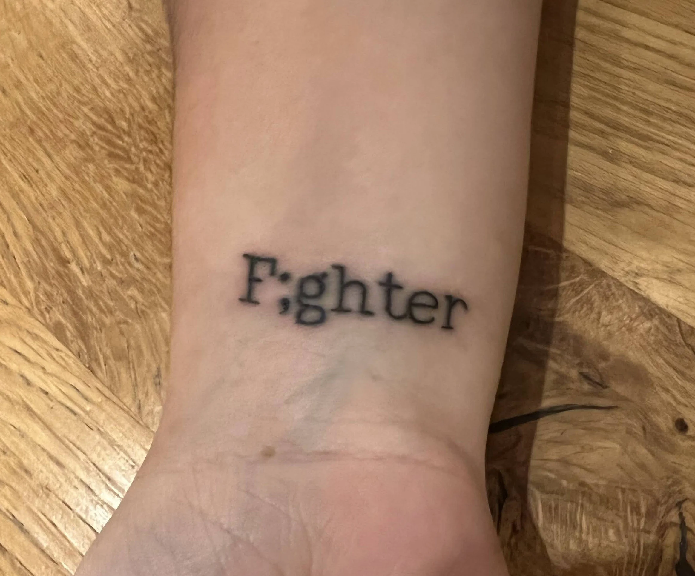 Close-up of an inner wrist with a black tattoo reading “F;ghter,” with a semicolon replacing the “i” in “Fighter,” against a wooden background.