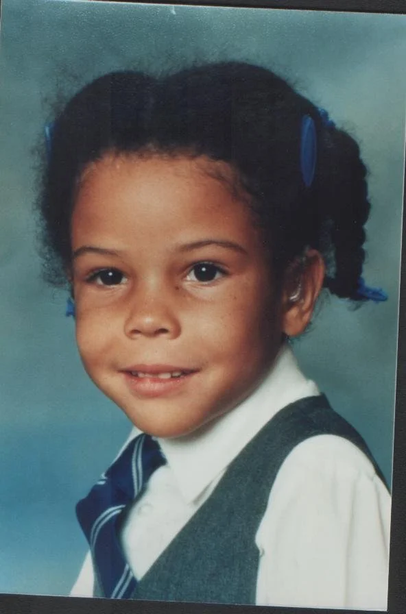 Photograph of a young child in a school portrait, facing the camera and smiling gently. The child wears a school uniform with a shirt, tie, and jumper, and their hair is styled in two sections with clips.