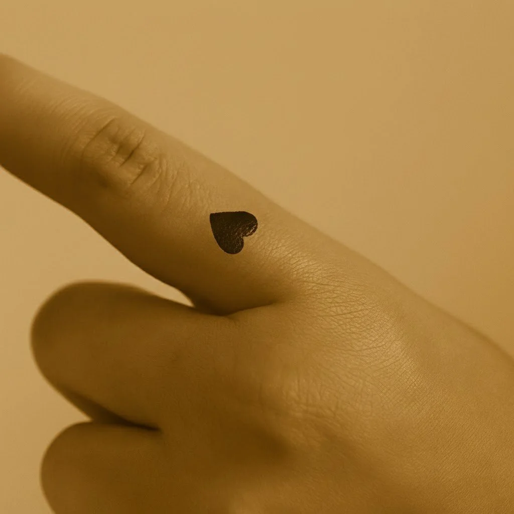 Photograph of a small black heart tattoo on the side of the left index finger. The image has a warm vintage sepia overlay, with the heart angled correctly to face upward toward the fingernail.