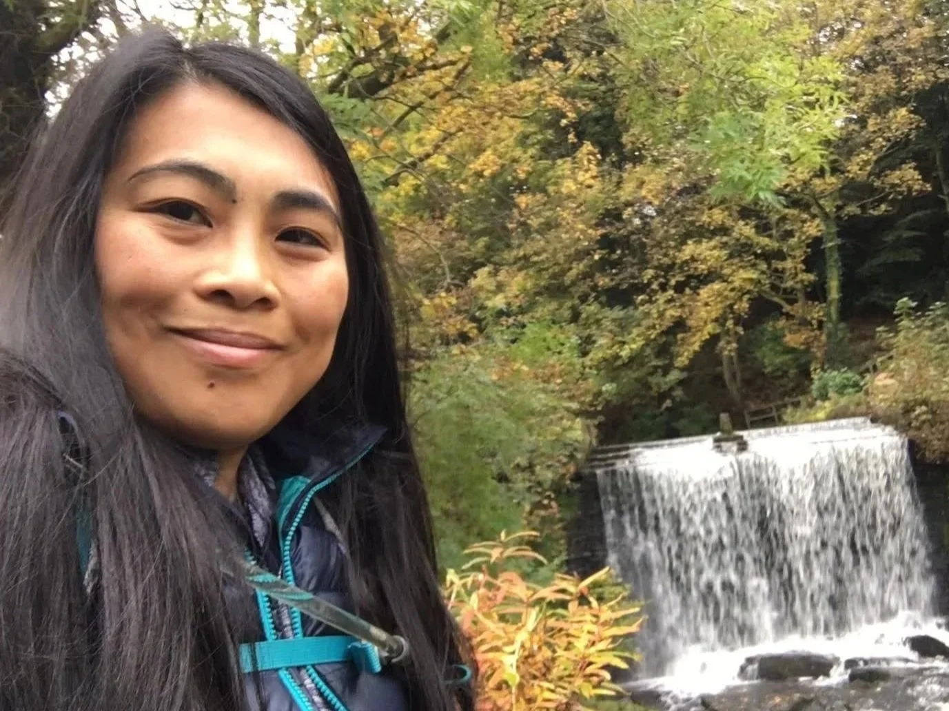 Woman with long black hair standing and taking a selfie with a waterfall
