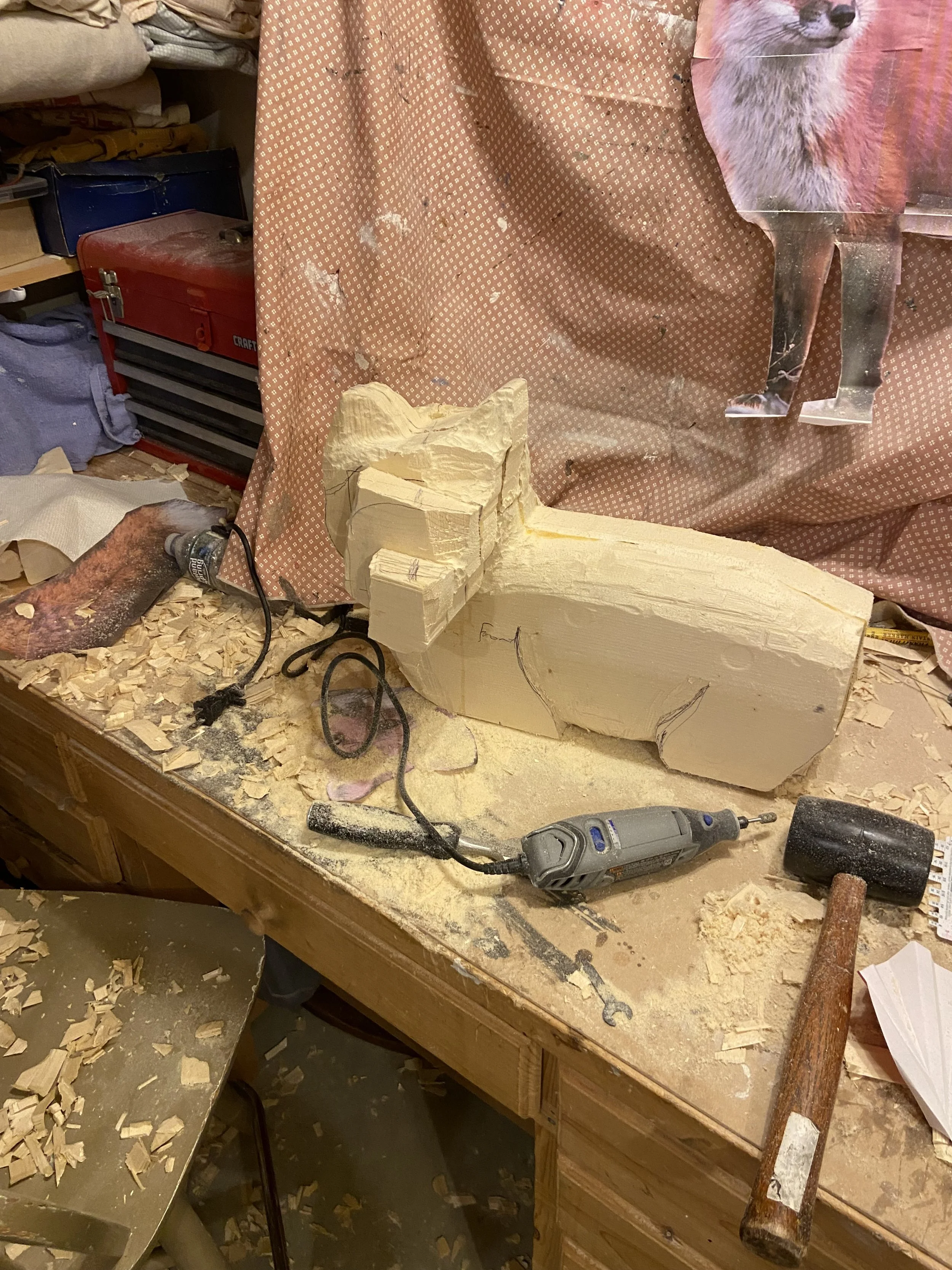 A woodworking workspace with a large piece of wood being carved, surrounded by wood chisels, a saber saw, a mallet, and wood shavings on the table and floor. There is a backdrop with a pink cloth and a poster of a dog.