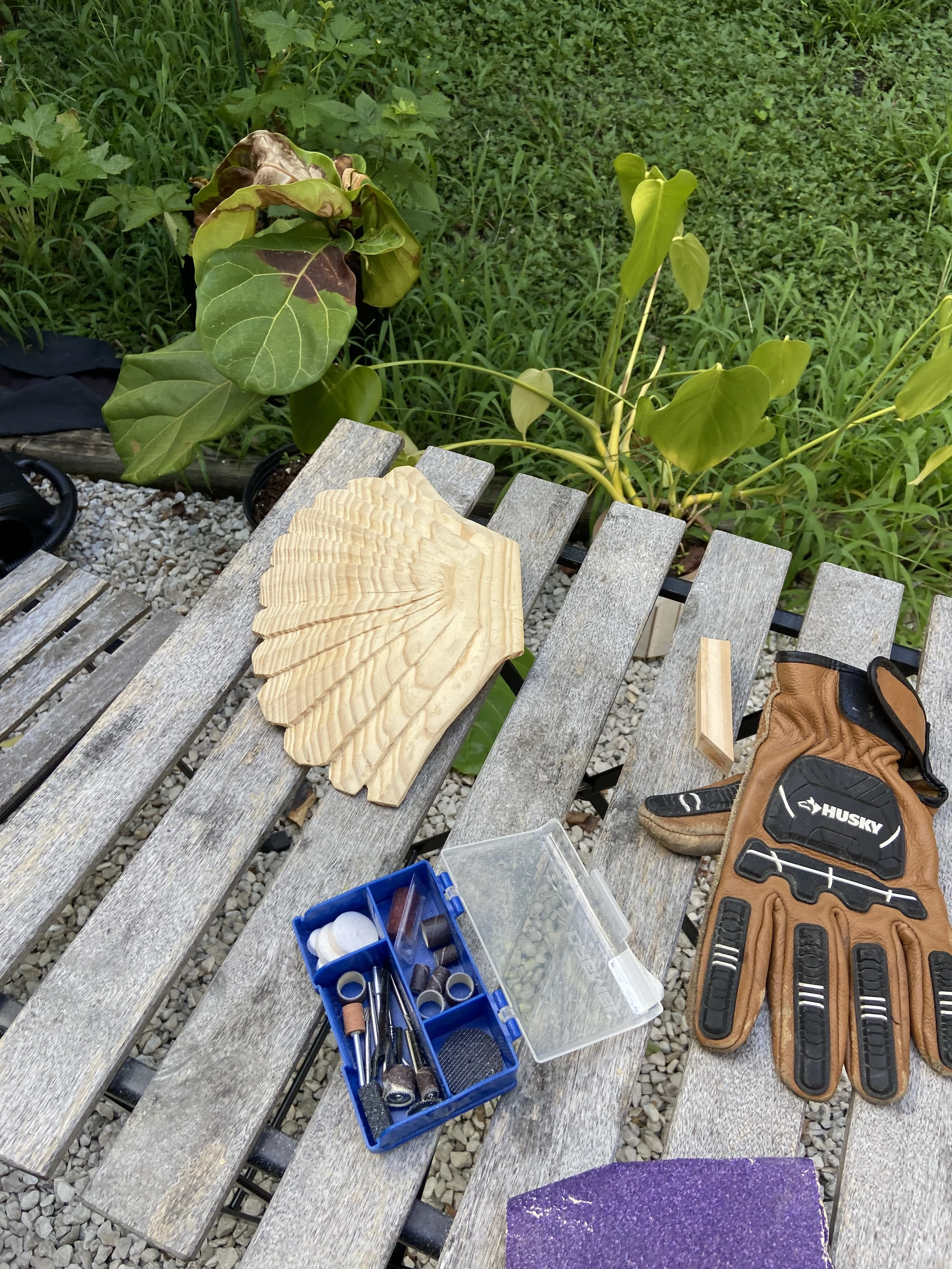 Wood carving tools, a partially carved wooden piece shaped like a leaf, a glove, and a small block of wood on a weathered outdoor bench with green plants in the background.
