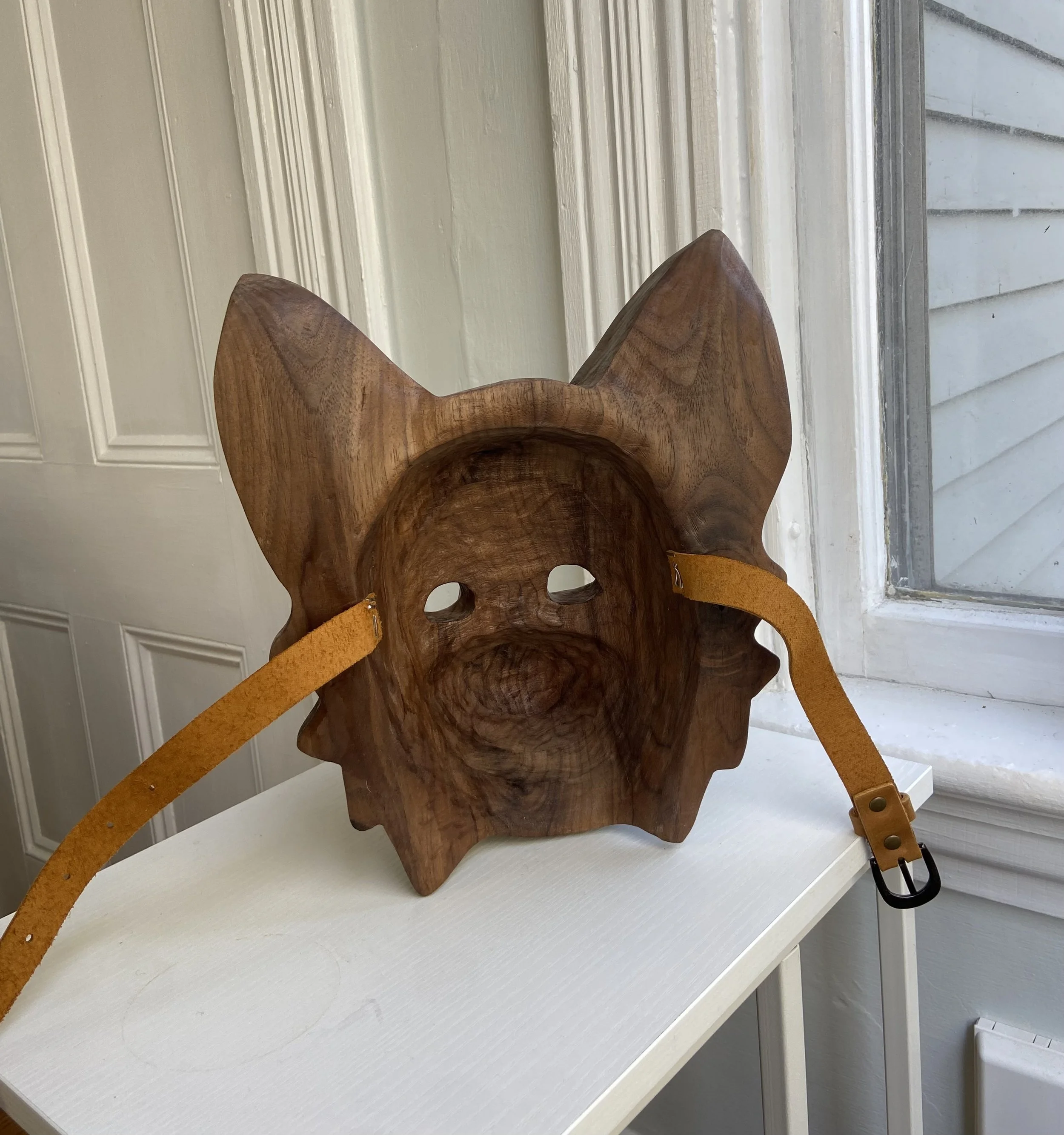 Wooden mask resembling a dog's face with large ears and cut-out eyes, hanging on a white shelf near a window.