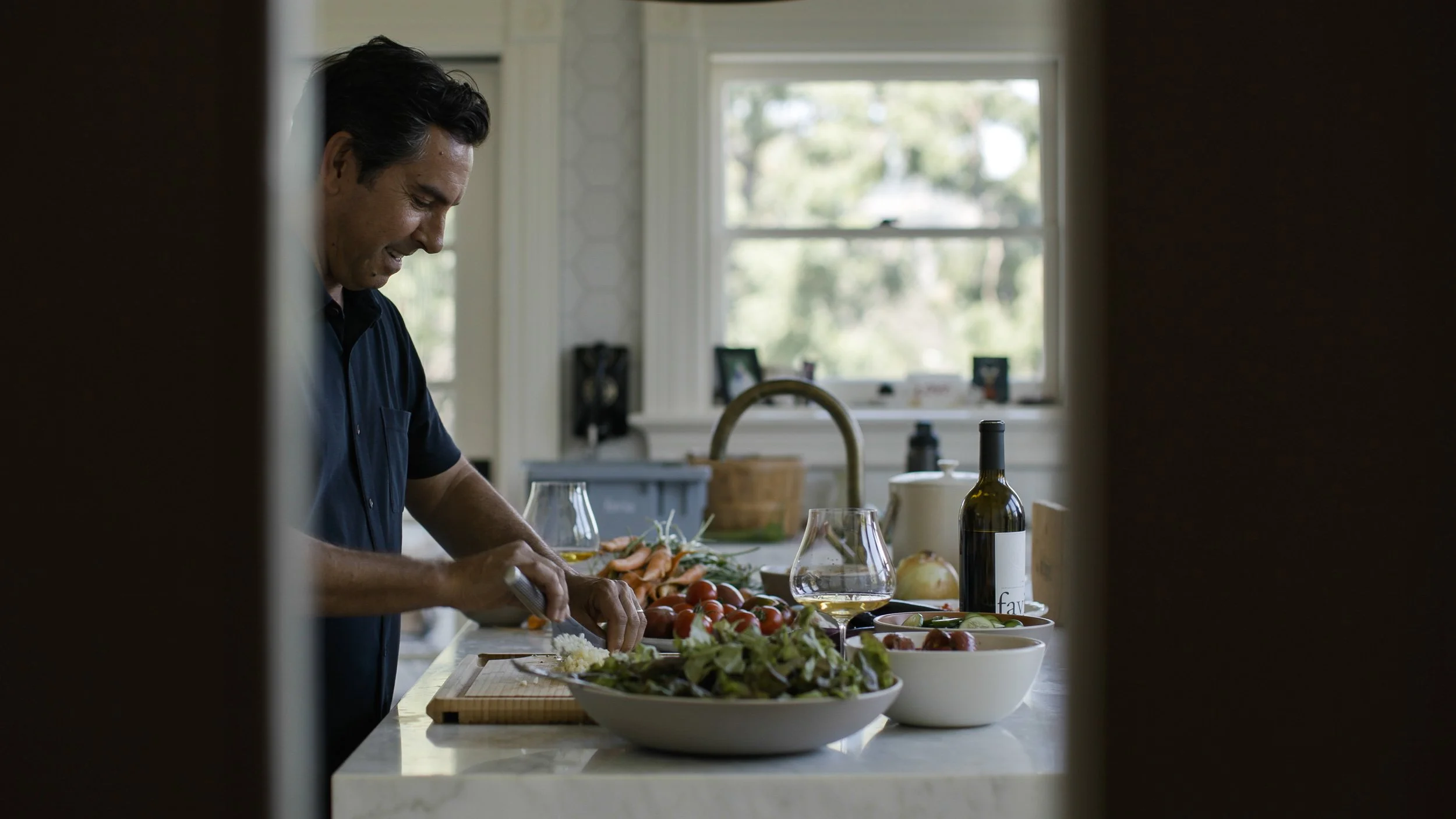 A man preparing salad in kitchen, viewed through a partial frame, with fresh vegetables, wine, and kitchenware on the countertop.