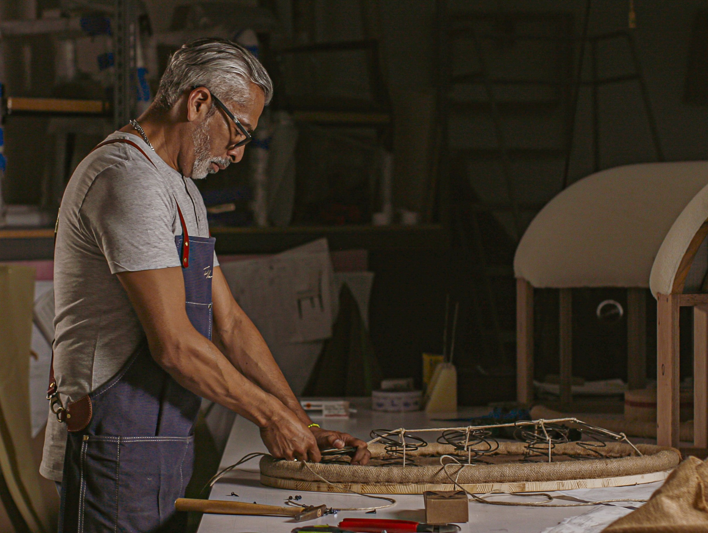 An older man with gray hair, glasses, and a gray beard working on a craft project at a workbench in a workshop, surrounded by tools and materials.