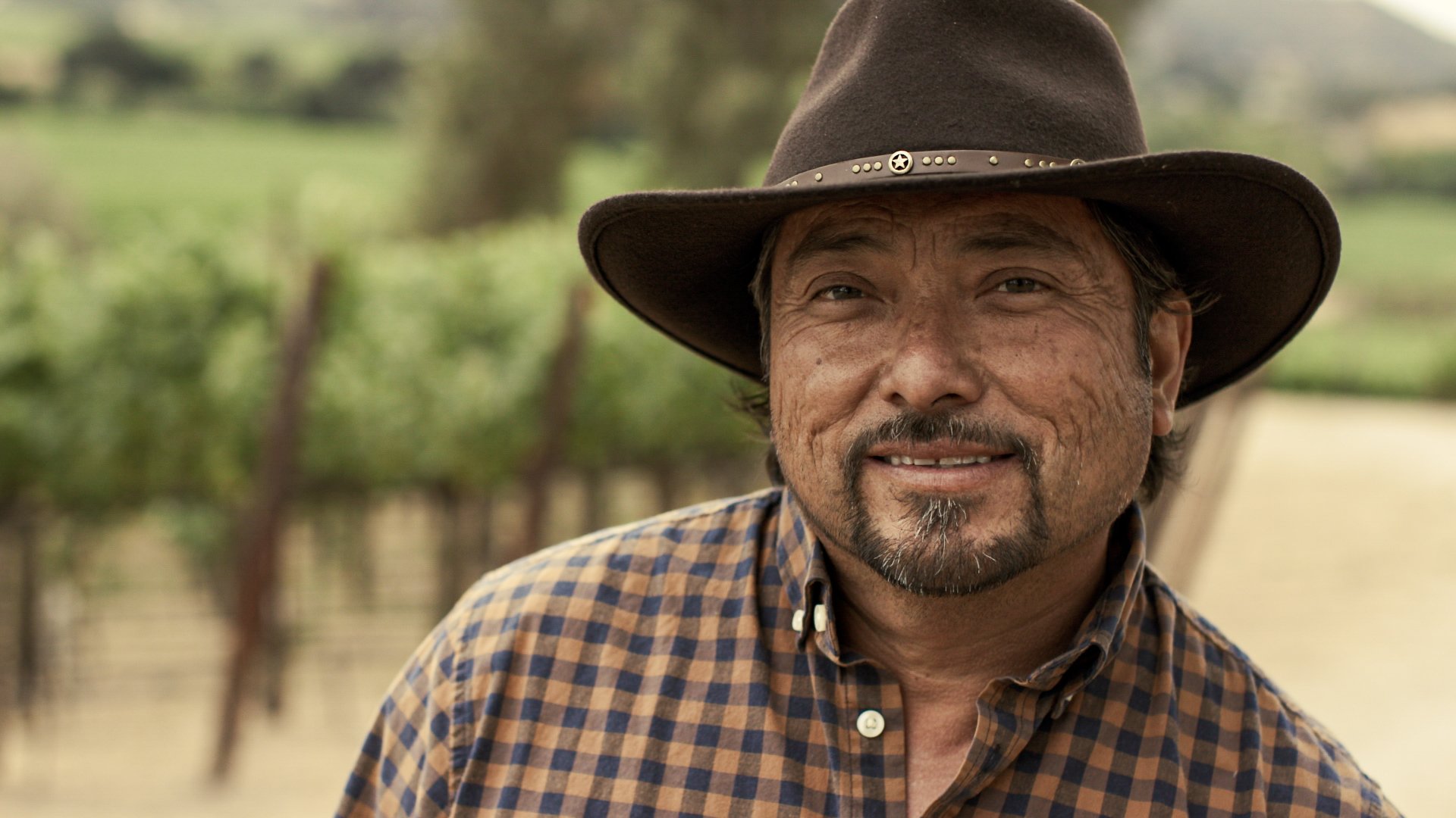 A middle-aged man with a beard smiling outdoors, wearing a brown wide-brimmed hat and a brown checkered shirt, with a vineyard in the background.