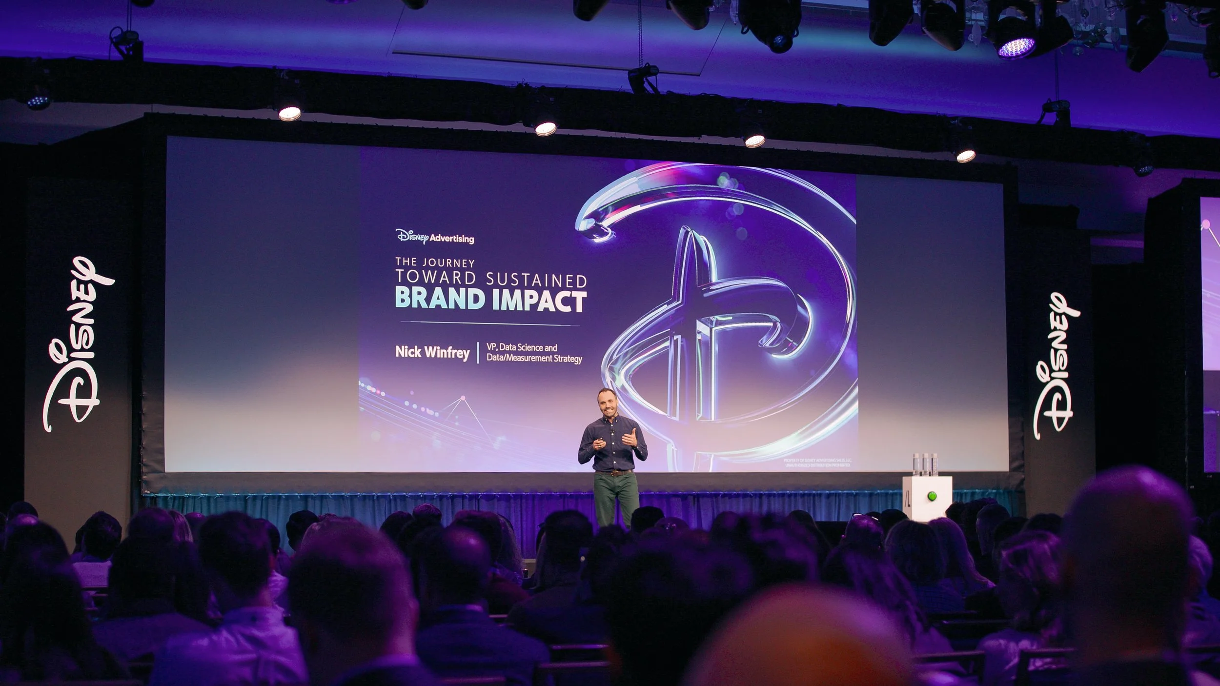 A man on stage giving a presentation at a Disney Advertising event, with a slide in the background titled 'The Journey Toward Sustained Brand Impact' and Disney logos on the sides.