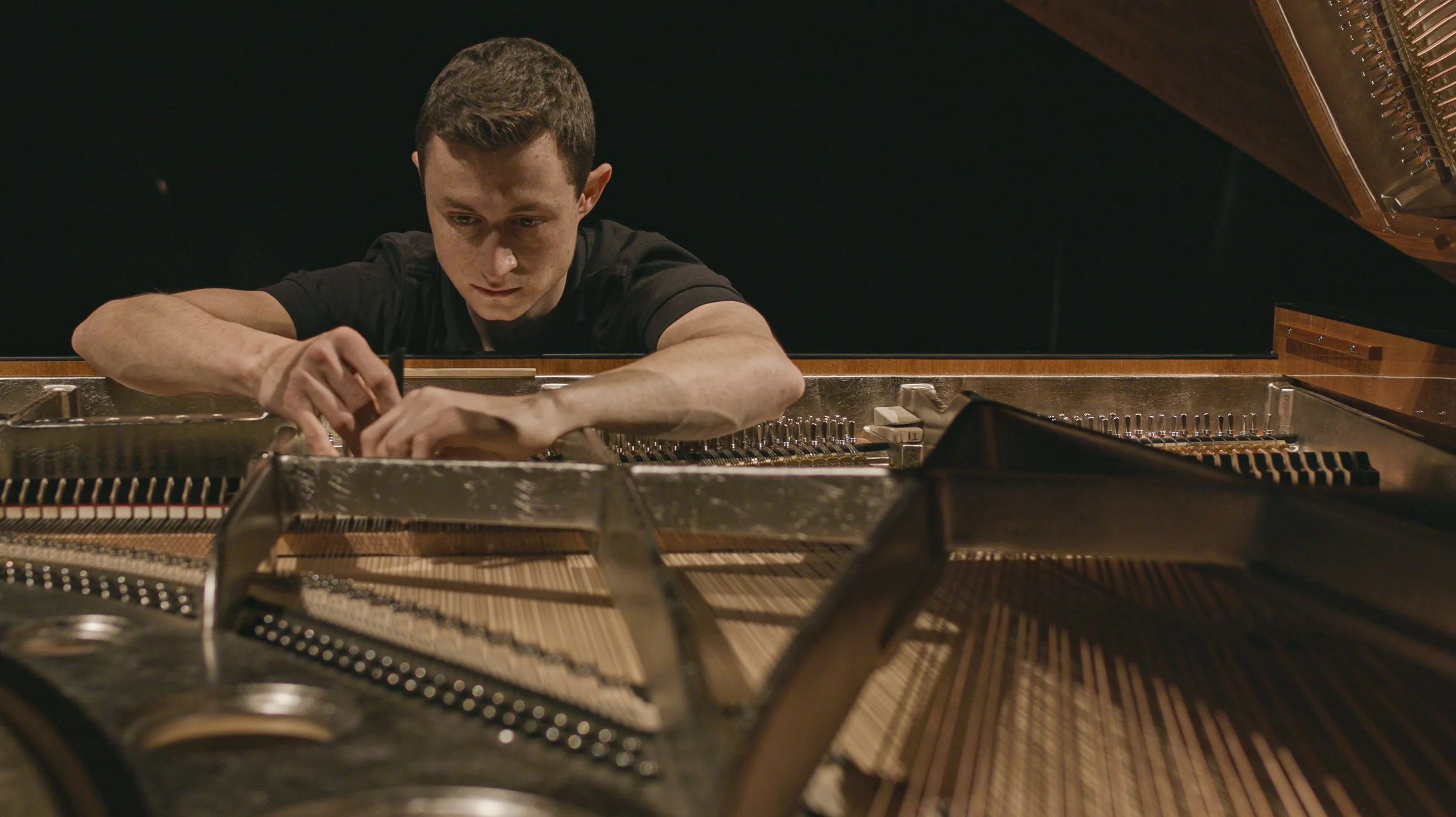A young man fixing or tuning a grand piano from inside, with the lid open showing the strings and tuning pins.