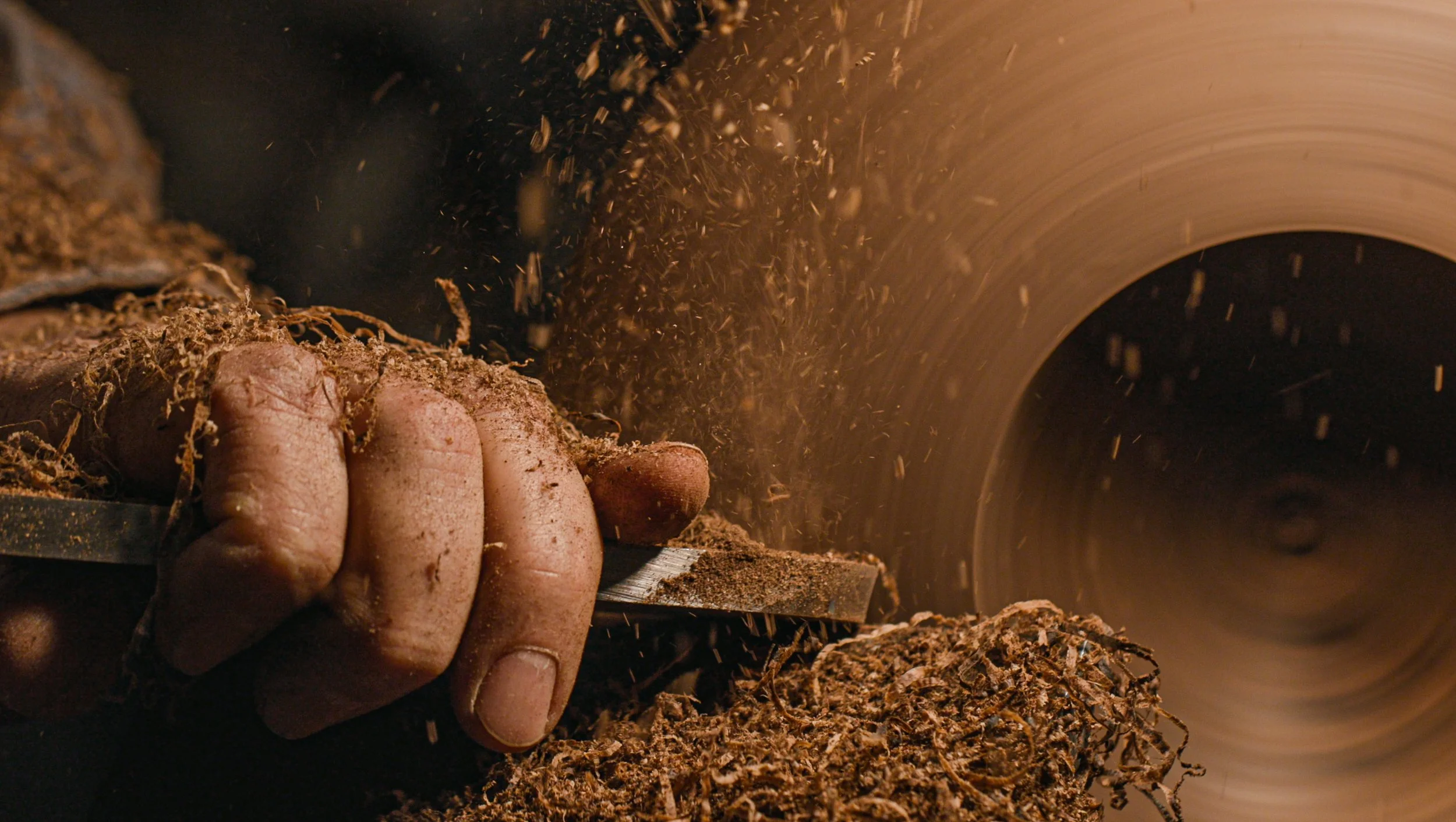 Close-up of a hand holding a tool cutting through a block of wood on a spinning saw blade, with sawdust and wood shavings flying in the air.