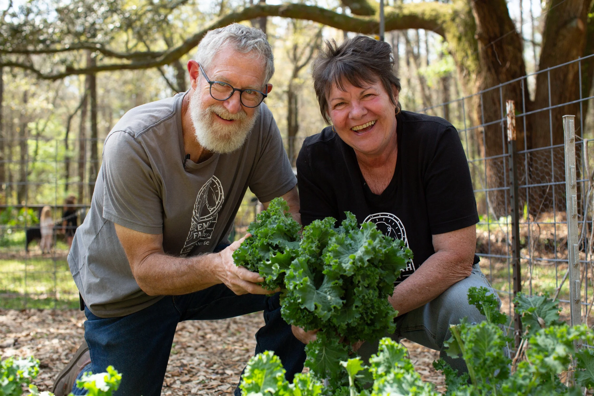 Older man and woman smiling in a garden, holding and showing a bunch of leafy green lettuce together, with trees and a fence in the background.