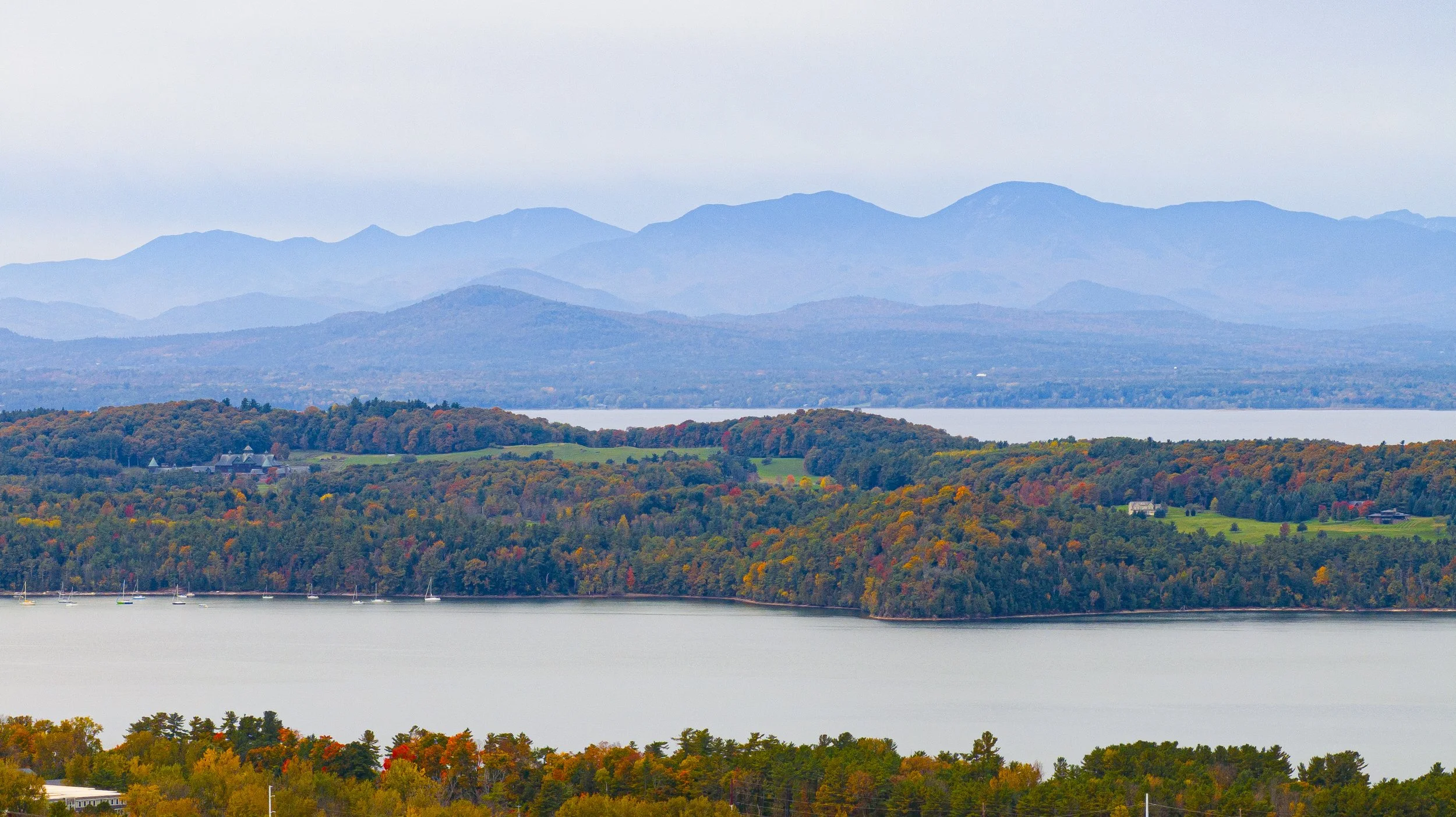 Scenic view of a large lake surrounded by forested hills with colorful fall foliage, and a mountain range in the distance under a cloudy sky.