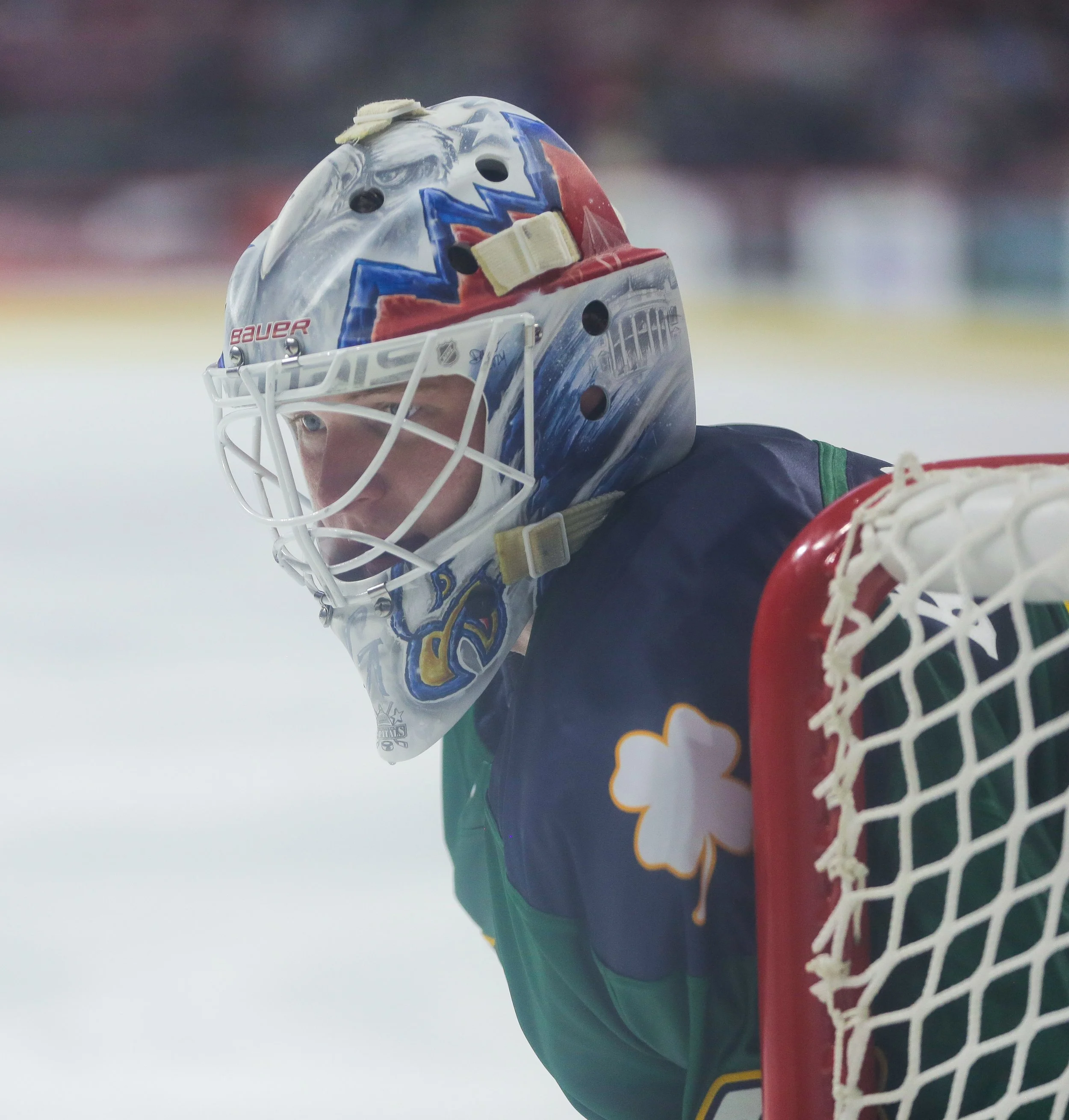 A young hockey goalie wearing a custom helmet and jersey, standing in front of the goal net on ice rink.