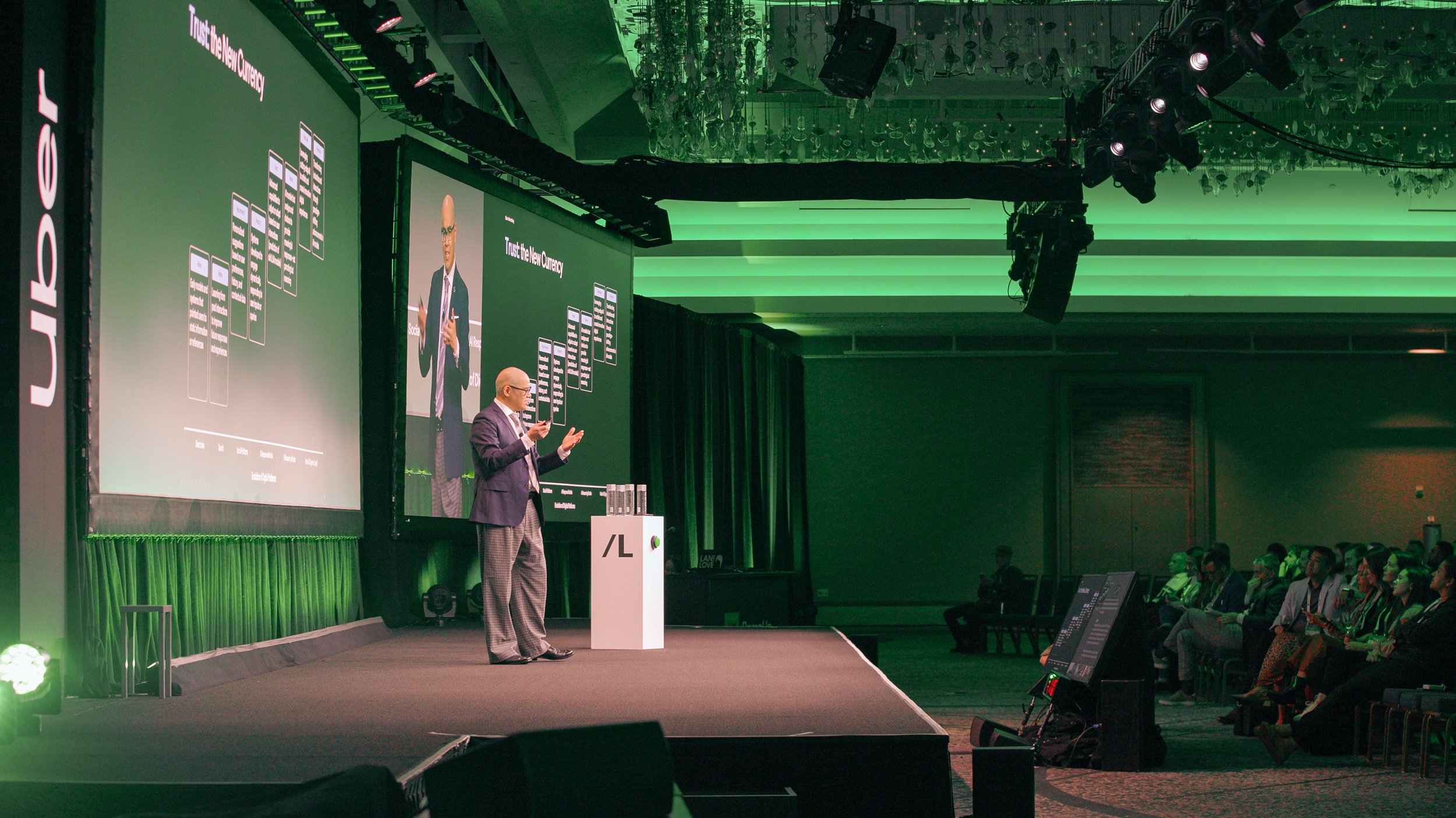 A man giving a presentation on stage at a conference with large screens behind him displaying a chart titled 'Trust the New Currency.' The audience is seated in front, with some taking notes or viewing the presentation.