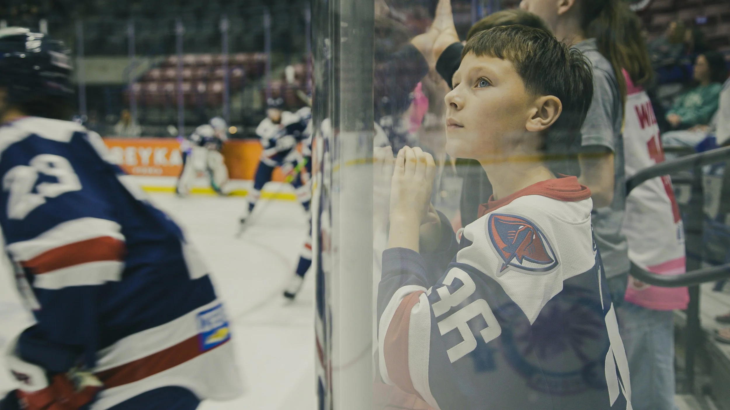 A young boy wearing a hockey jersey watching a hockey game through the glass at the rink.