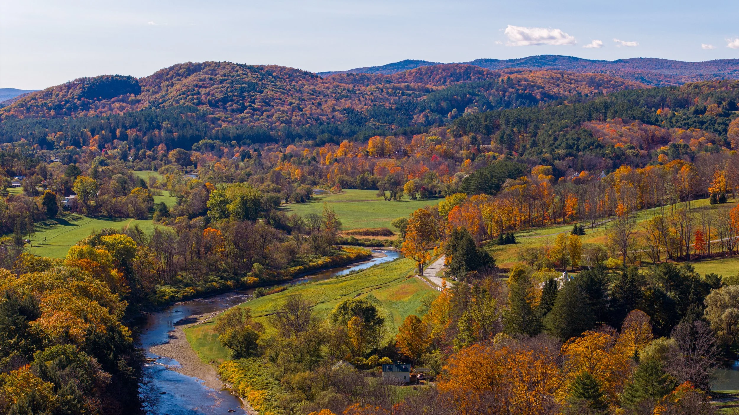 A fall landscape with a winding river, colorful trees, green fields, and rolling hills in the distance.