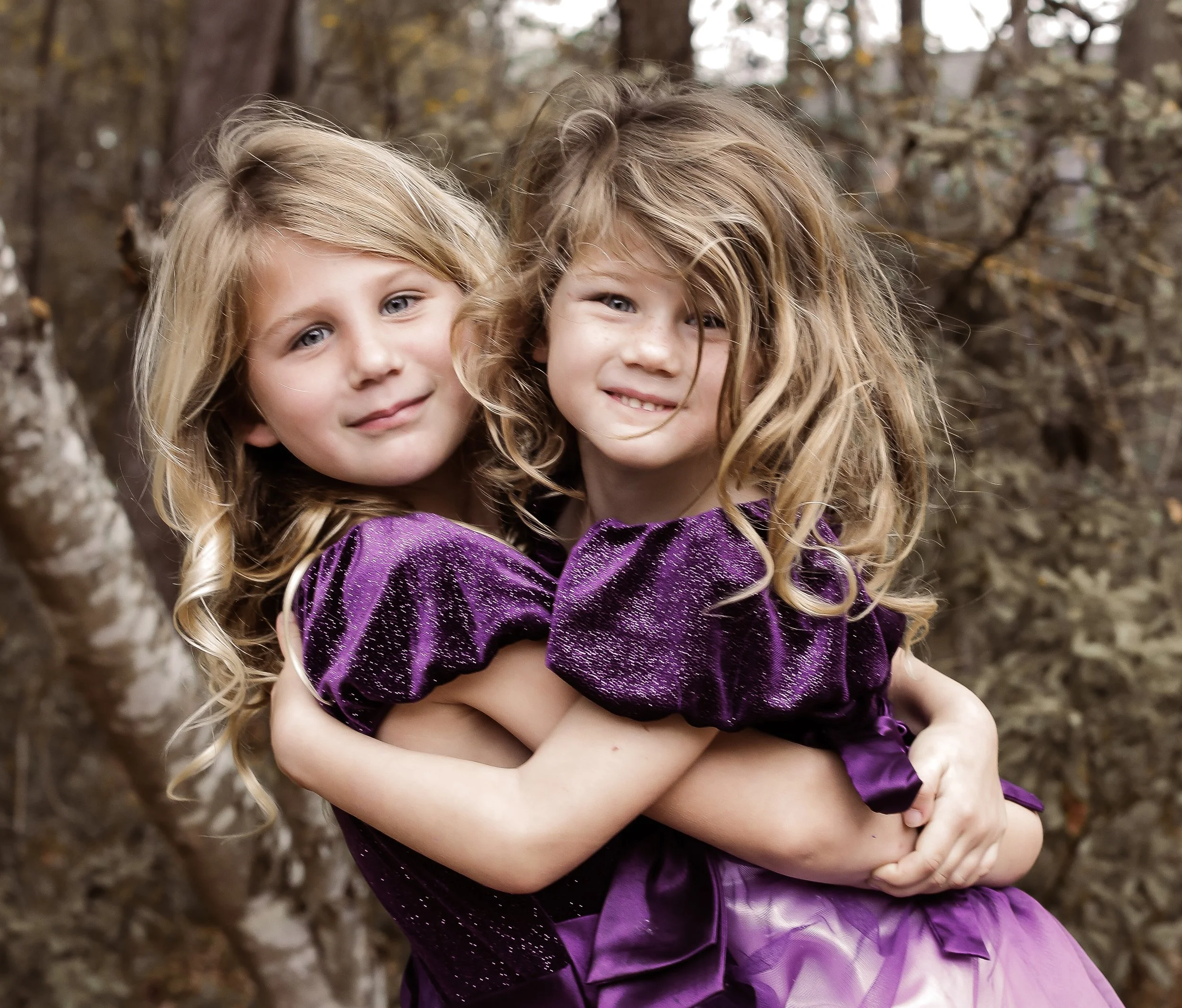 Two young girls with curly blonde hair embrace each other outdoors in front of trees, wearing matching purple velvet and tulle dresses.