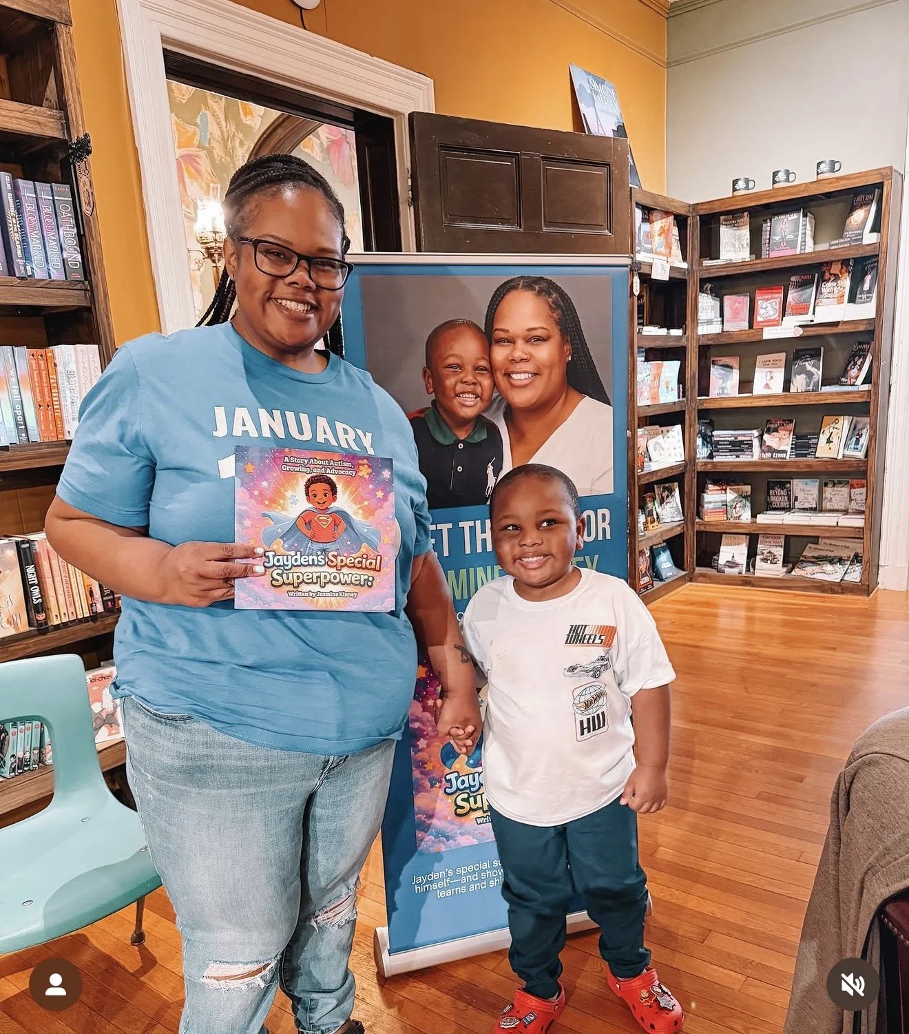 A woman holding a children's book titled 'Jayden's Special Superpower', standing next to a young boy in a bookstore. The woman has glasses and braided hair, and is wearing a blue shirt and distressed jeans. There is a poster of a mother and child in 