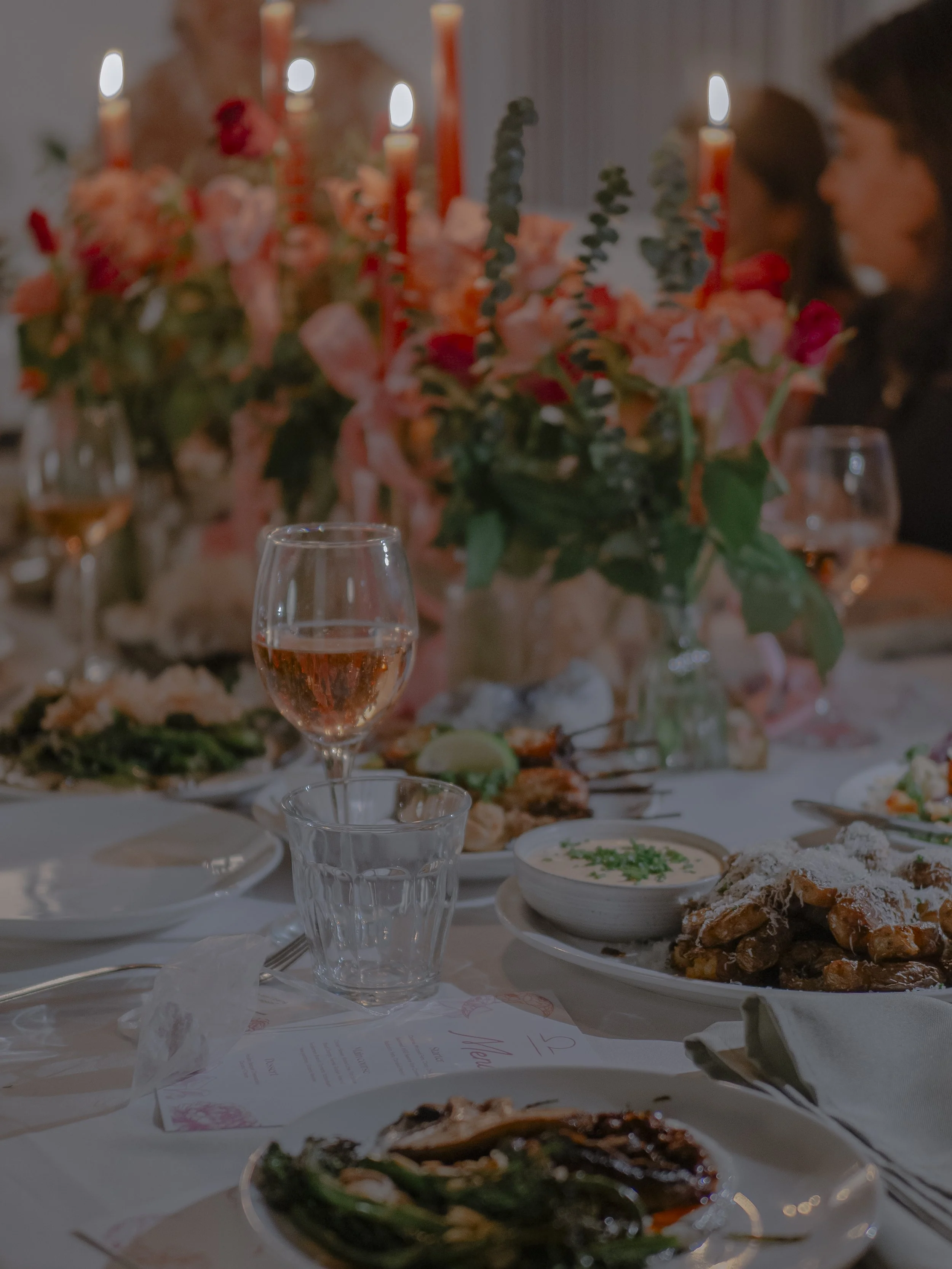 A supperclub, a beautiful dining table with a large floral centerpiece, lit candles, glasses of rosé wine, and various dishes including salad, sauce, and roasted vegetables.