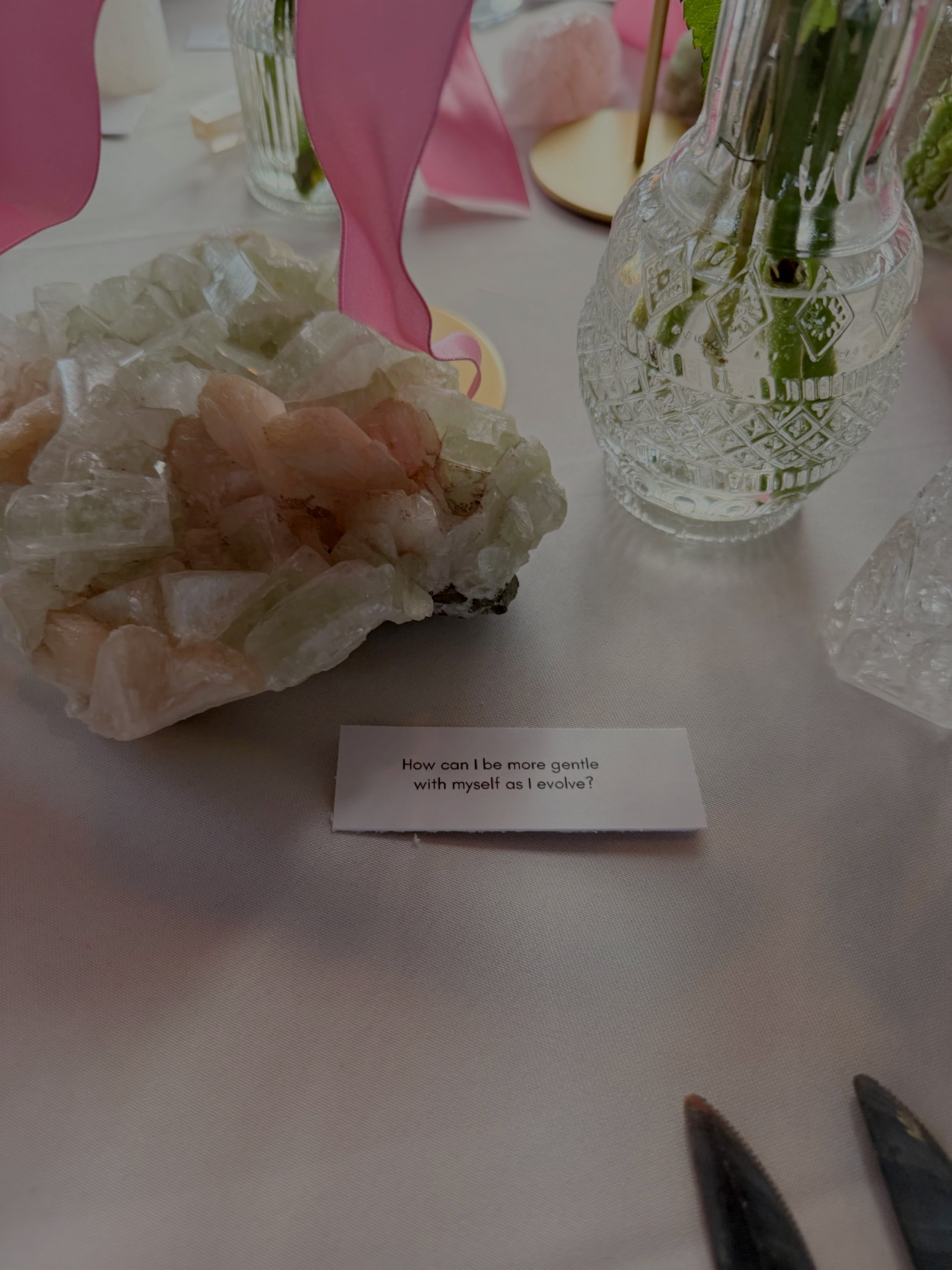 Table setting with a large pink and cream-colored mineral specimen, a clear patterned glass vase with flowers, pink napkins, and a printed note reading 'How can I be more gentle with myself as I evolve?'