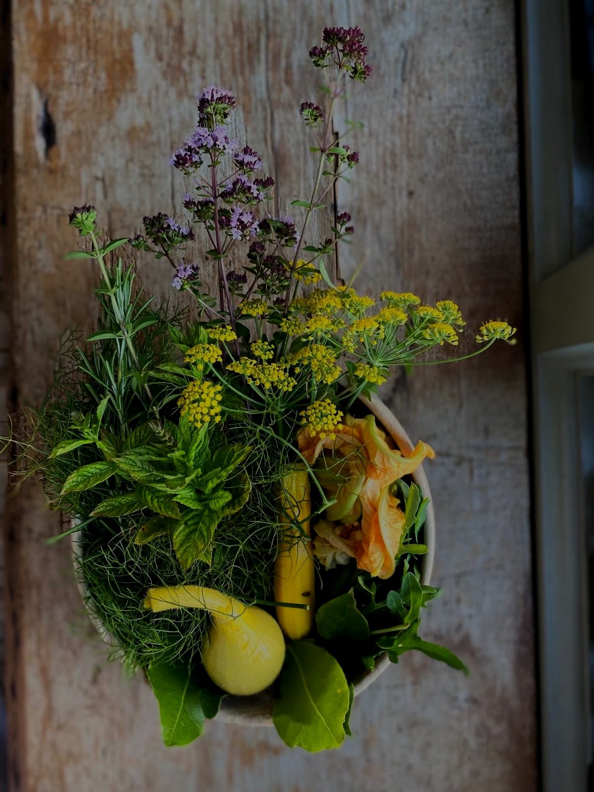 A decorative arrangement of flowers and produce in a white oval container on a wooden surface. The arrangement includes purple and yellow flowers, green leafy plants, a yellow zucchini, a yellow squash, and orange flowers.