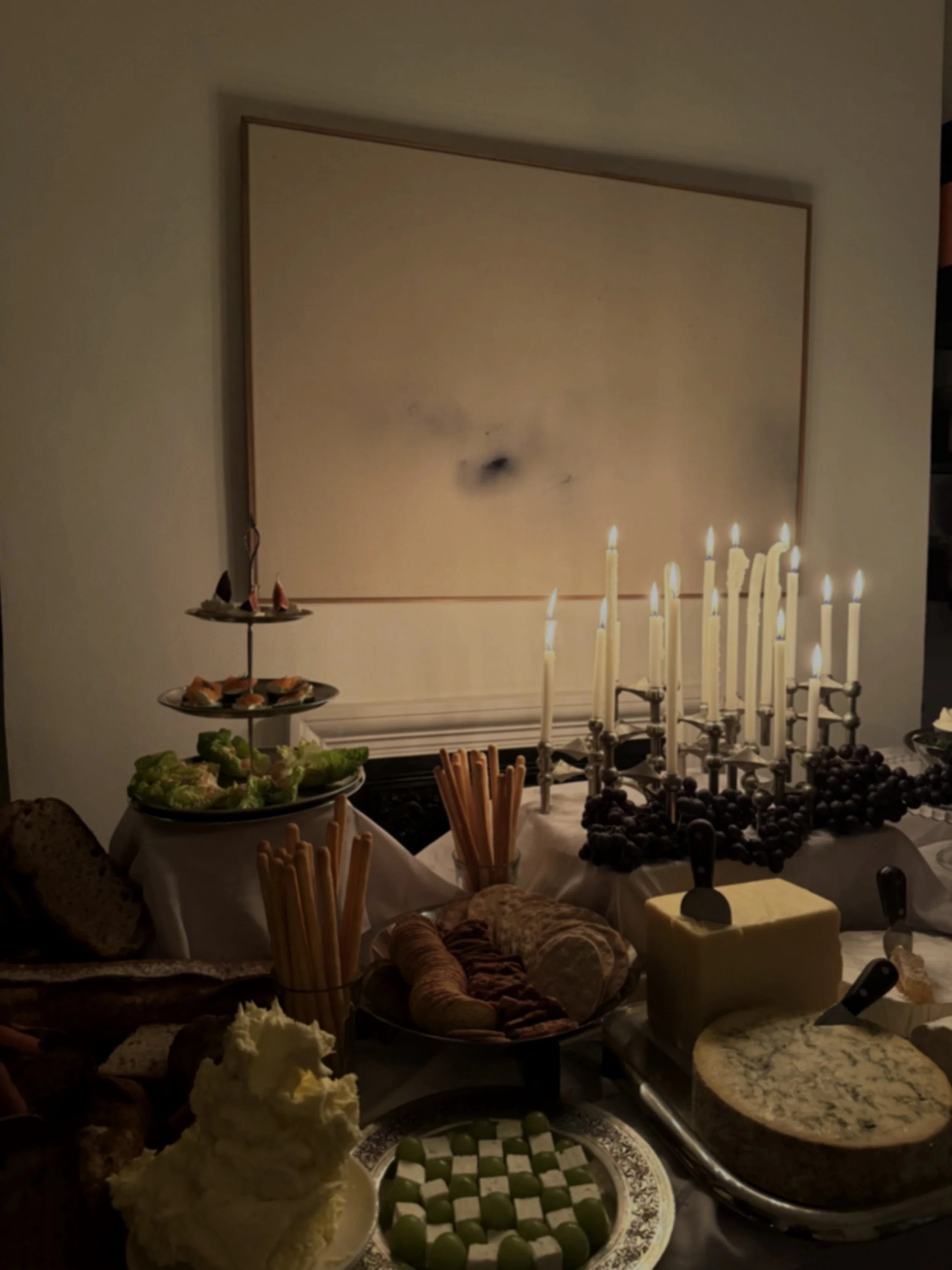 A table set with a variety of cheeses, bread, vegetables, and crackers, with lit candles and a large framed abstract painting in the background.