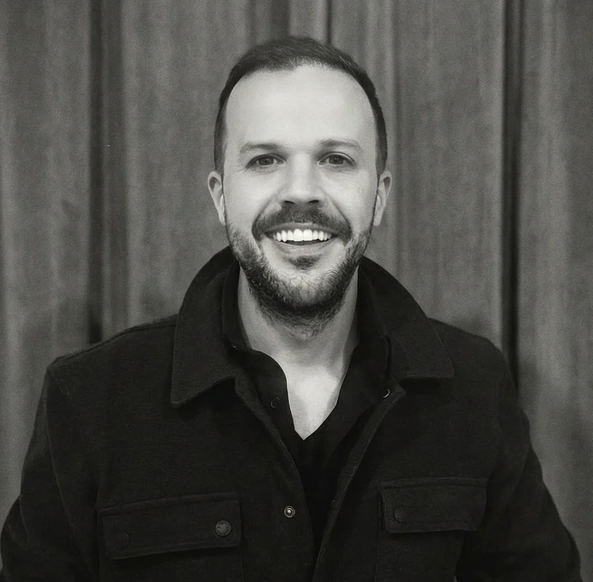 Black and white portrait of a smiling man with short hair and a beard, wearing a dark jacket and a shirt, standing in front of a wooden background.