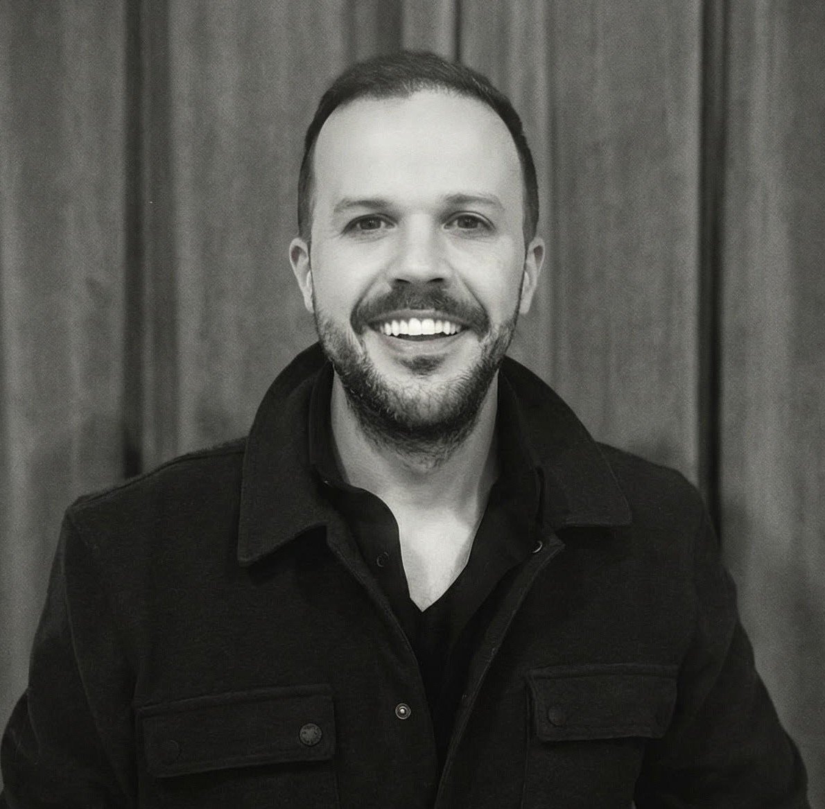 Black and white photo of smiling man with beard, wearing a dark jacket and shirt, standing in front of a wooden background.