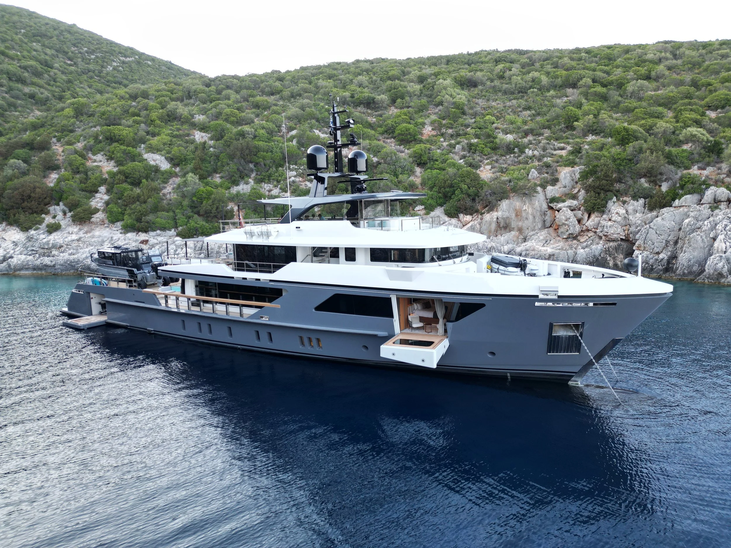 Luxury yacht anchored near rocky, green hillside with calm water.