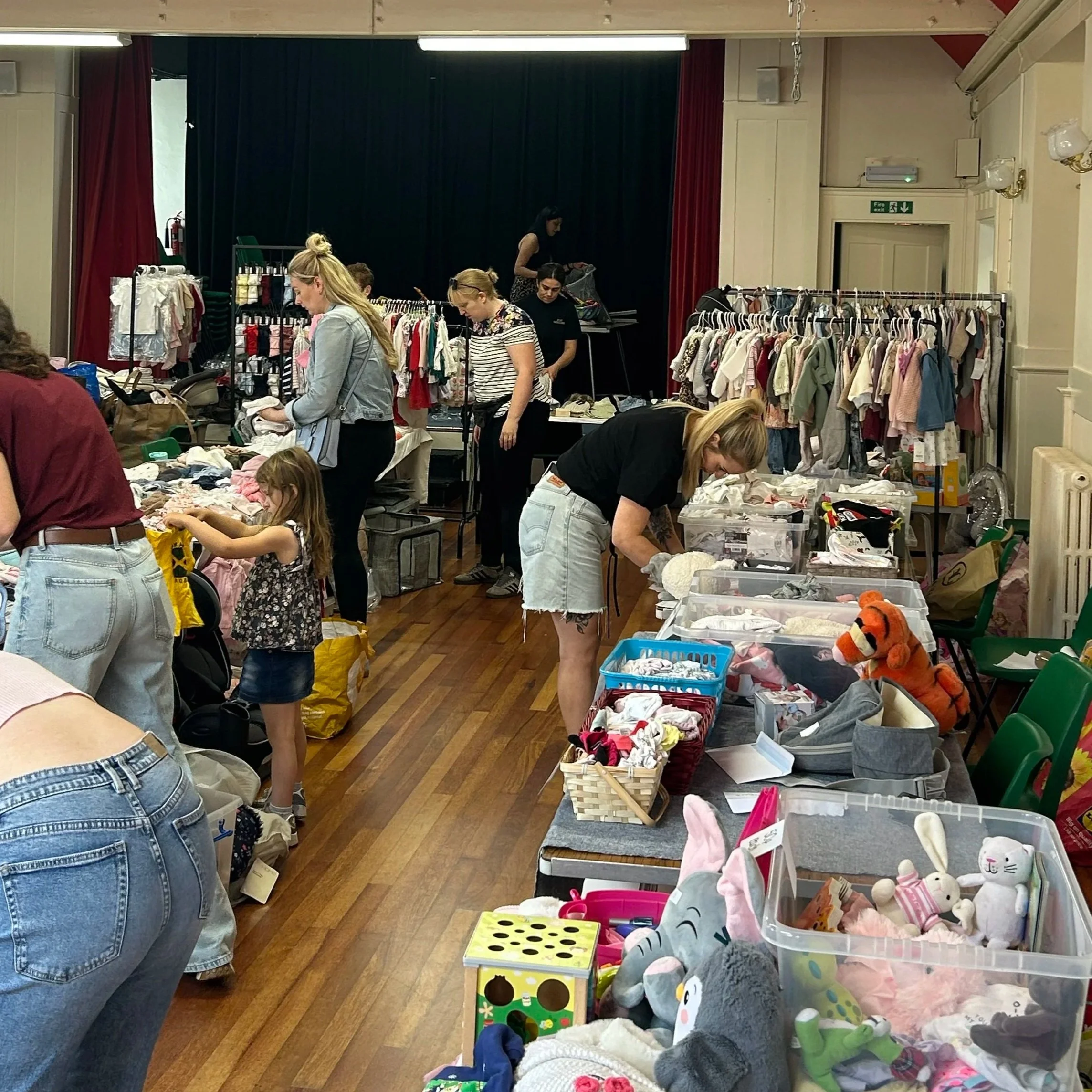 People browsing and shopping at a rummage sale or charity event with tables filled with toys, clothes, and donations inside a hall.