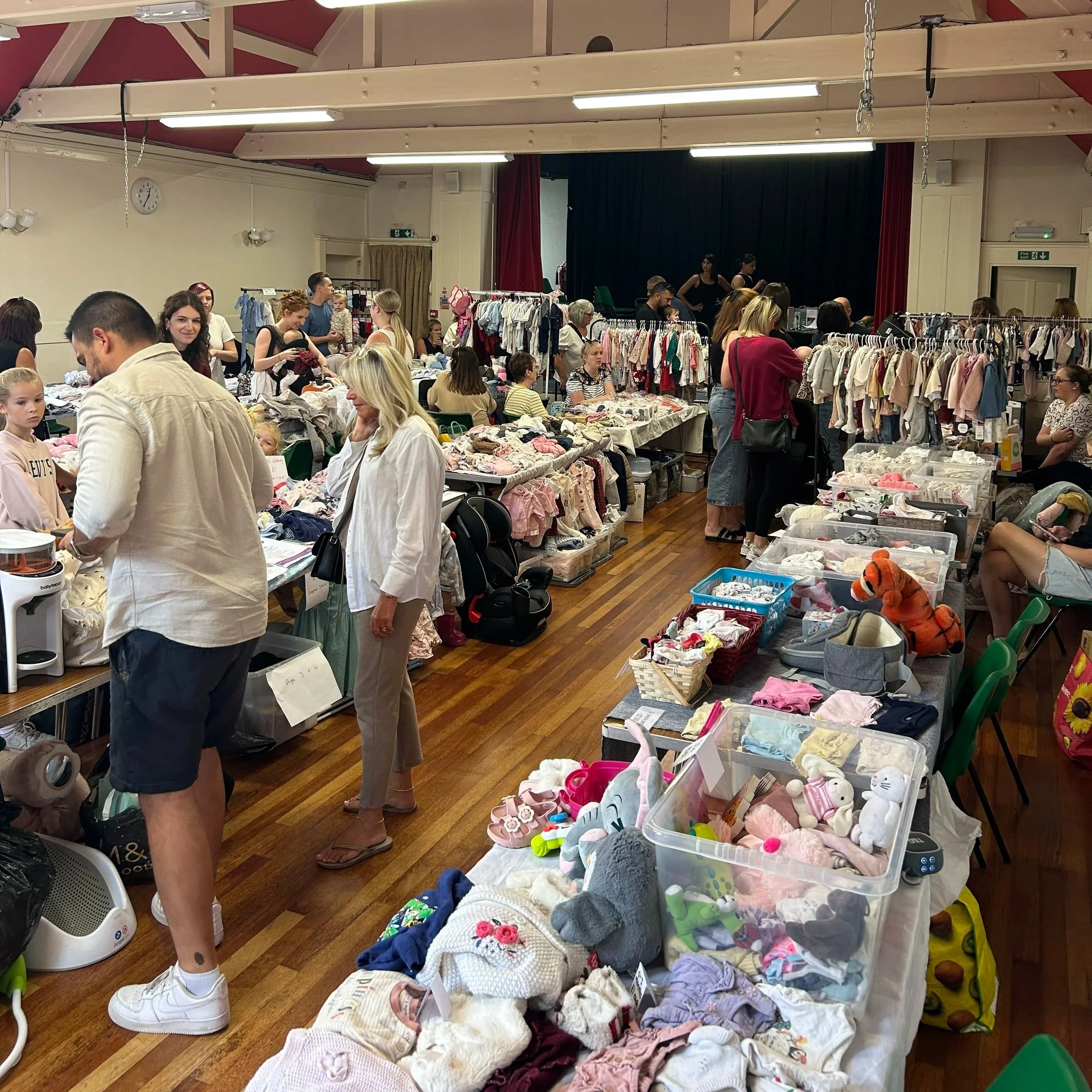 People browsing various items at a flea market or thrift sale inside a large hall with wooden flooring. Racks of children's clothing and tables with toys and stuffed animals are visible, and some individuals are seated and chatting.