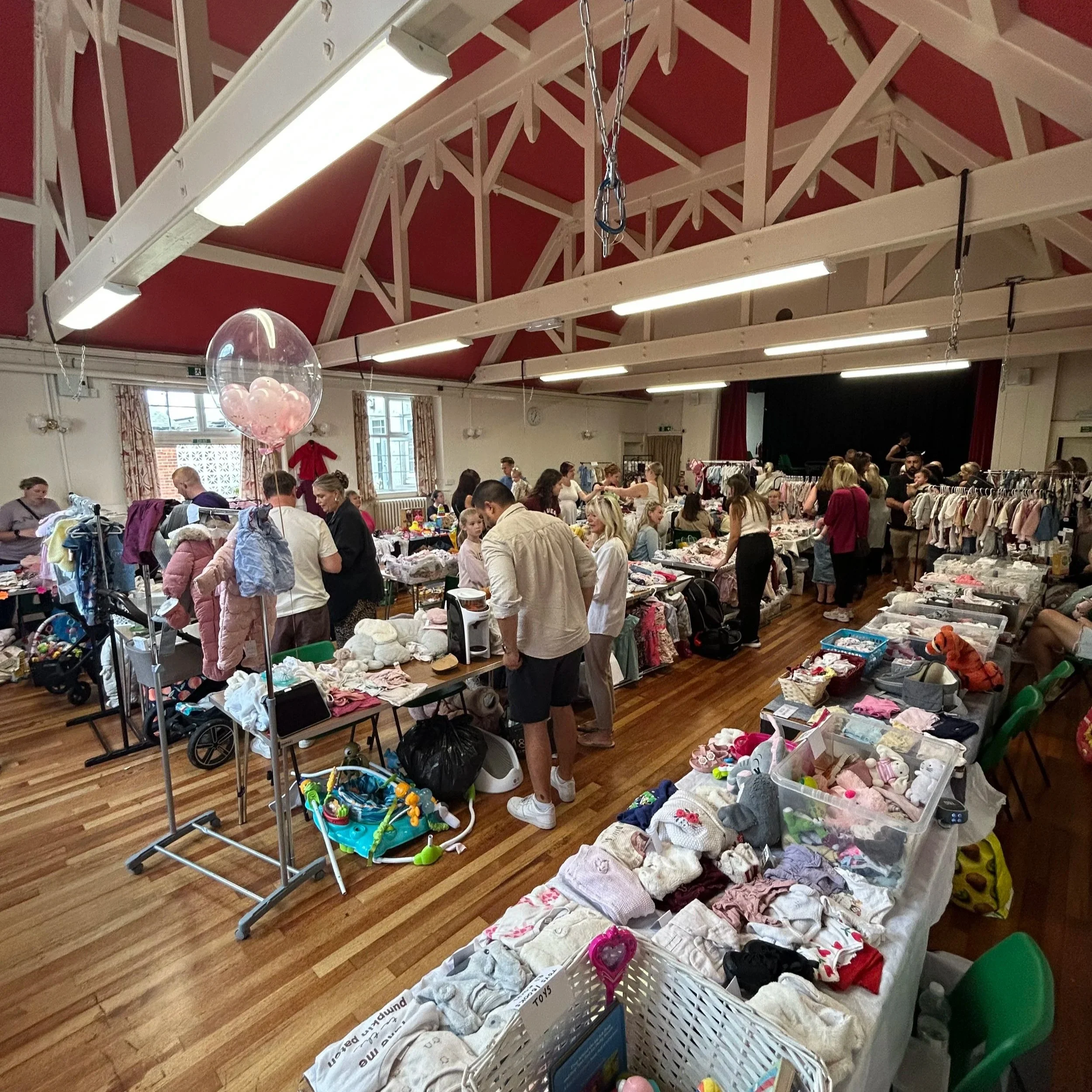 A crowded indoor clothing and toy sale event with tables filled with baby clothes, toys, and accessories. People browse and shop among racks of children's clothing, and the room features a high ceiling with exposed beams and fluorescent lighting.