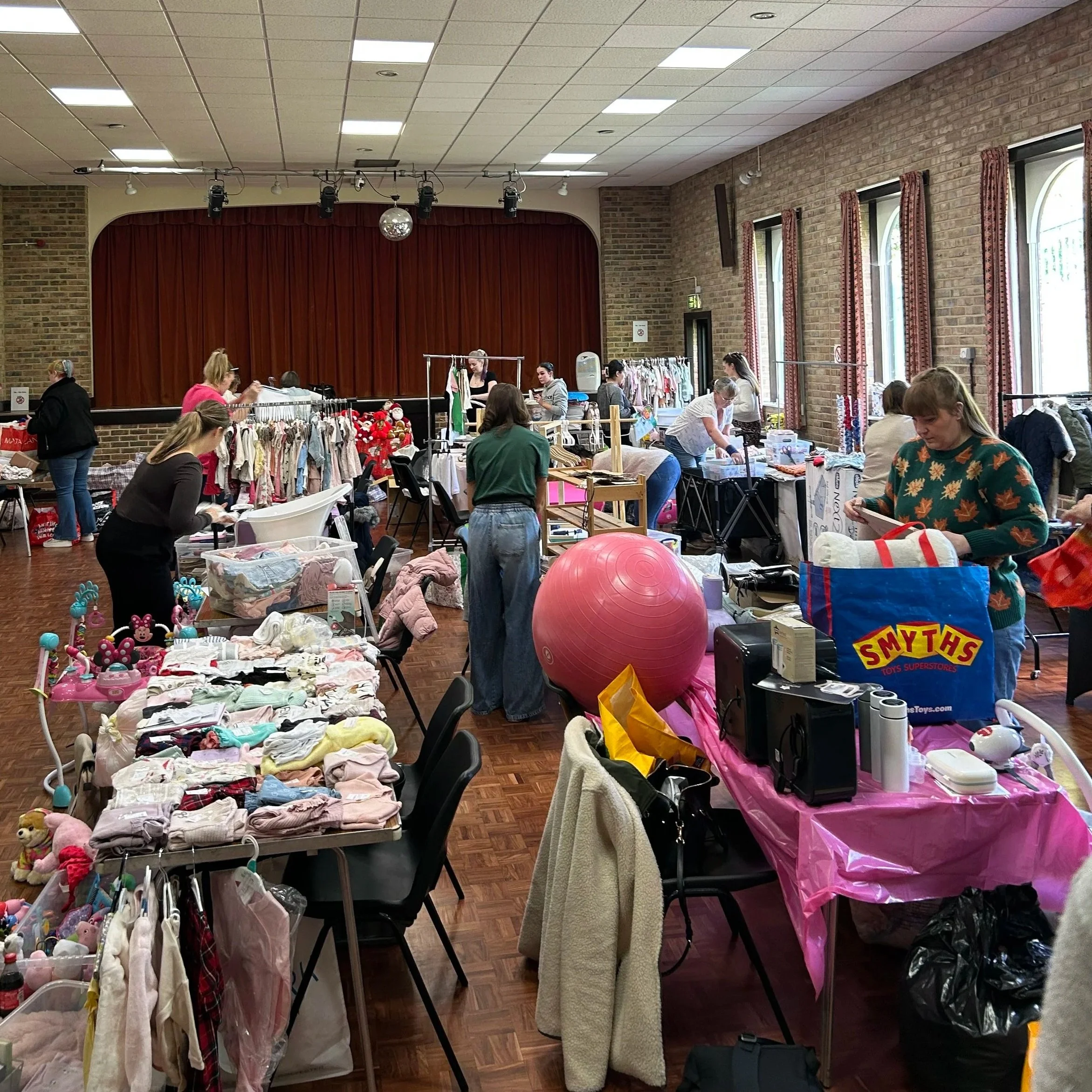 Inside a community hall with people shopping at various tables and clothing racks, and toys displayed on tables, with a stage with red curtains in the background.