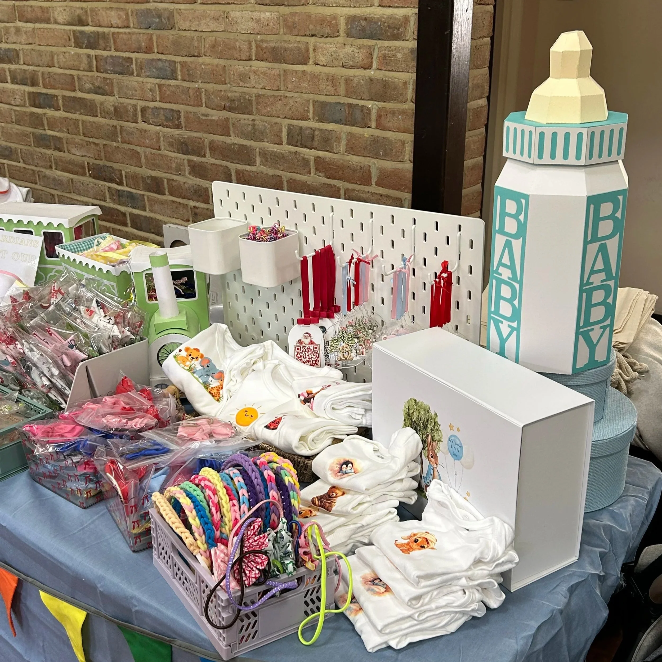 Table displaying baby-themed items such as diapers, clothing, headbands, and toys, including a large baby bottle-shaped gift box in the background, against a brick wall.