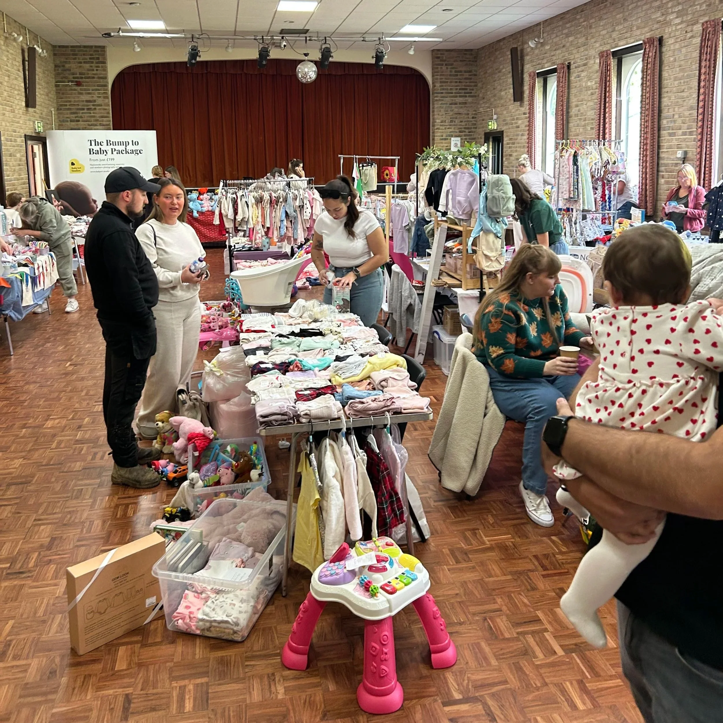 People shopping at a swap meet or indoor market with tables and racks filled with secondhand children's clothing, toys, and accessories. Several people are browsing and talking.