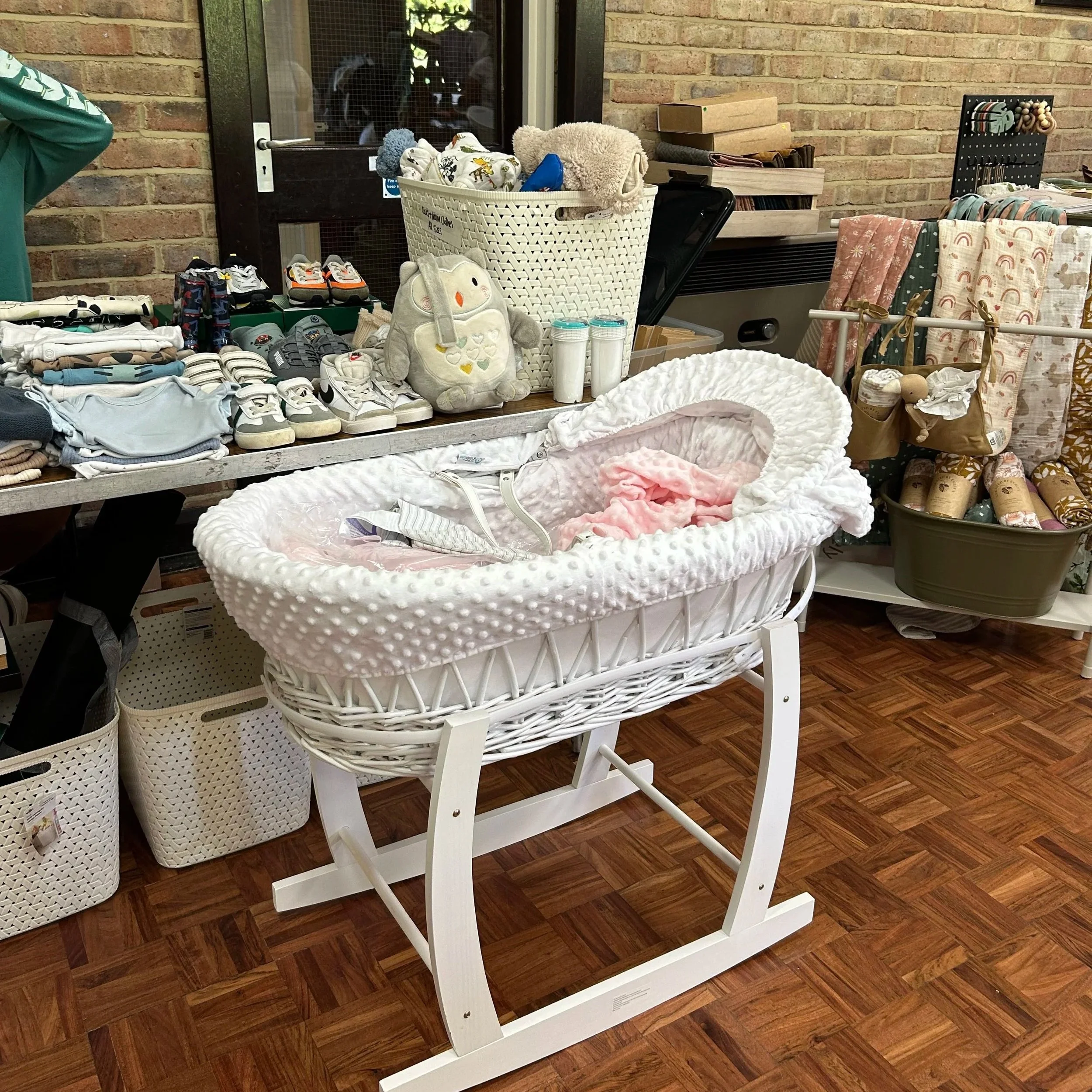 A white wicker bassinet with a pink blanket inside, displayed on a white stand at a thrift store or marketplace.