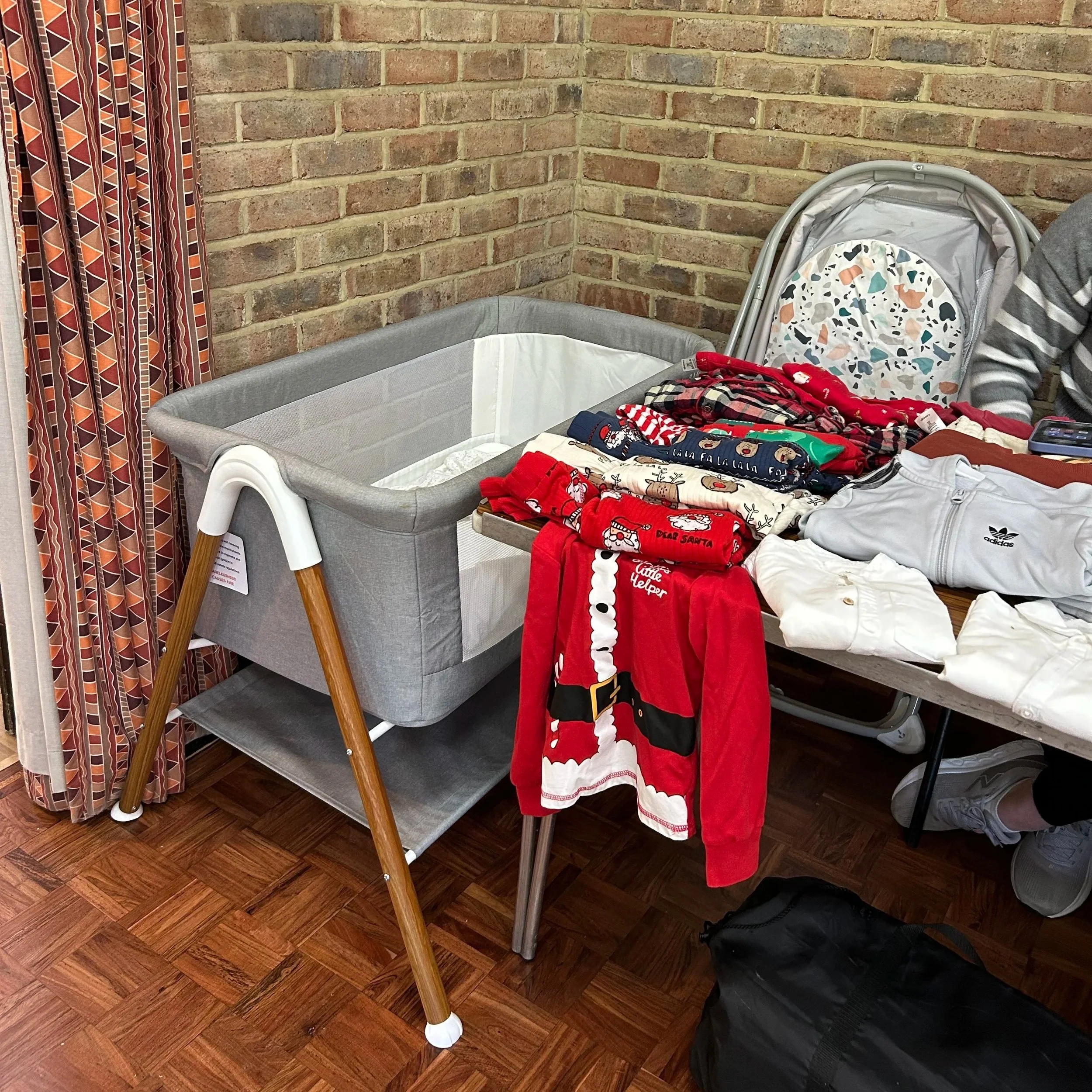 A changing table with a gray cushioned surface and a diaper storage under, next to a table filled with Christmas-themed clothing including Santa costumes, pajamas, and jackets. A baby stroller with colorful patterned fabric is behind the table, again