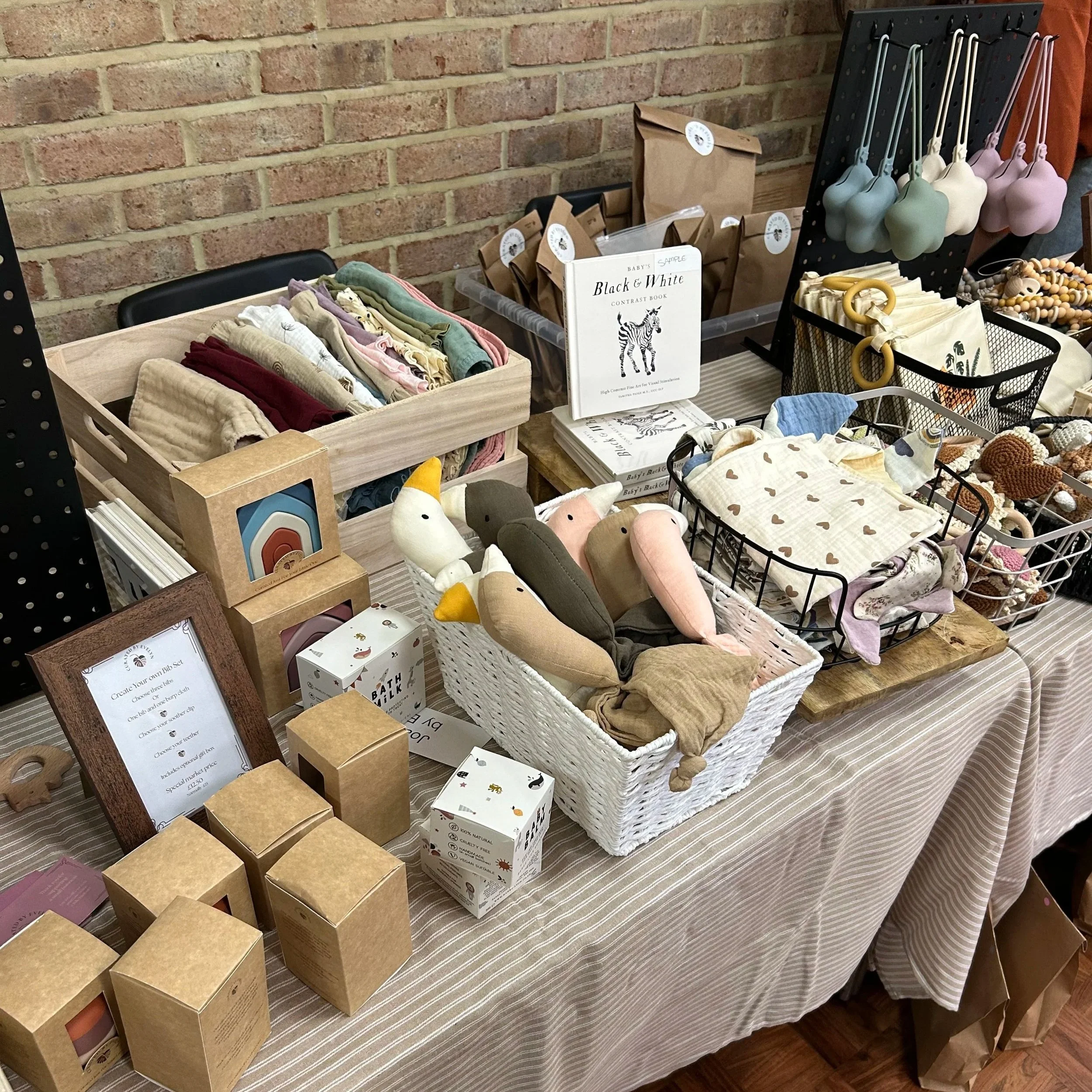 Display table with baby clothing, toys, and accessories arranged in baskets and containers against a brick wall.
