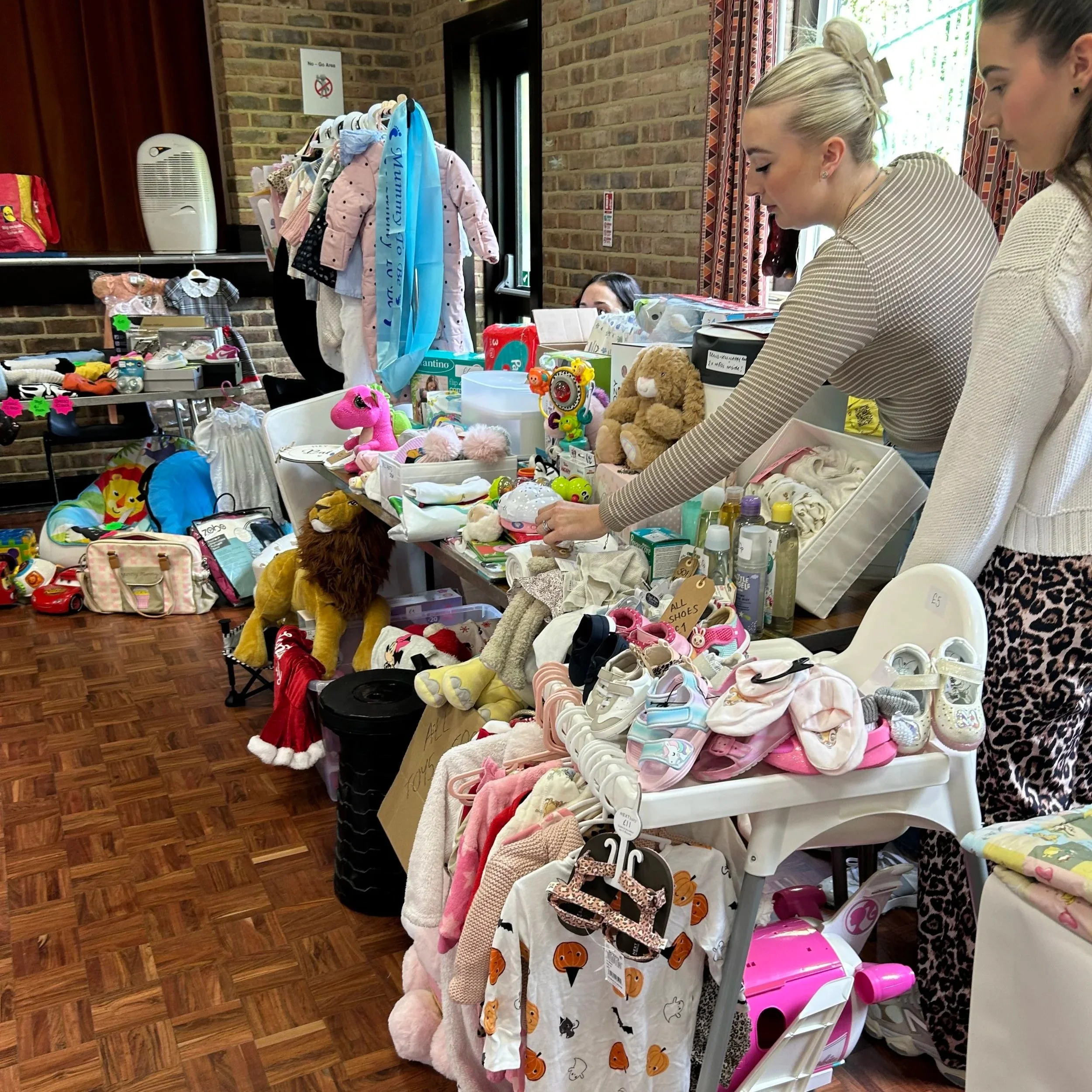 People at a flea market stall with toys, clothing, and baby items displayed on tables and racks inside a room with brick walls.