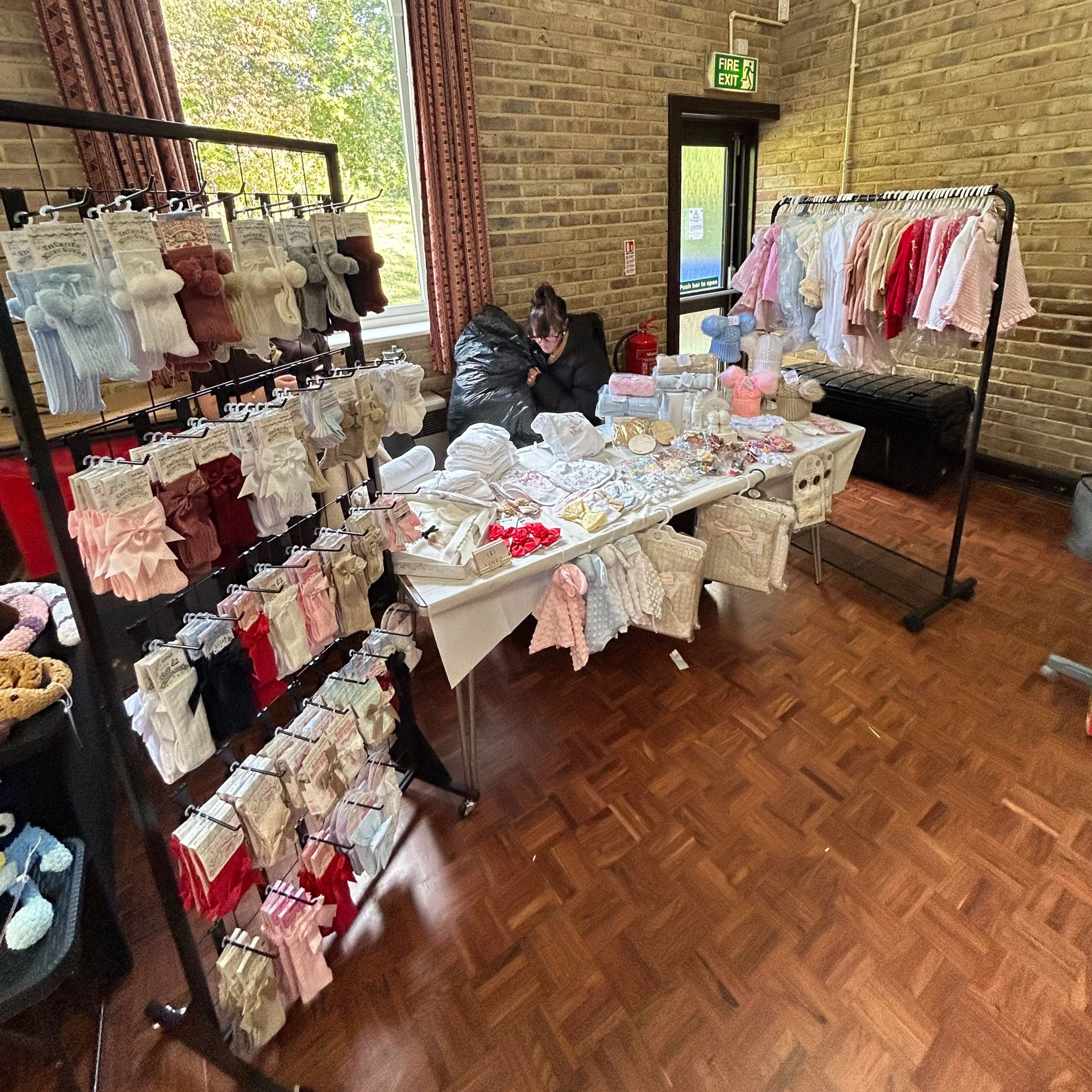 Indoor craft or gift market stall displaying knitted socks, headbands, and baby clothing on racks and tables with a person arranging items, brick walls, a window, and an exit door.